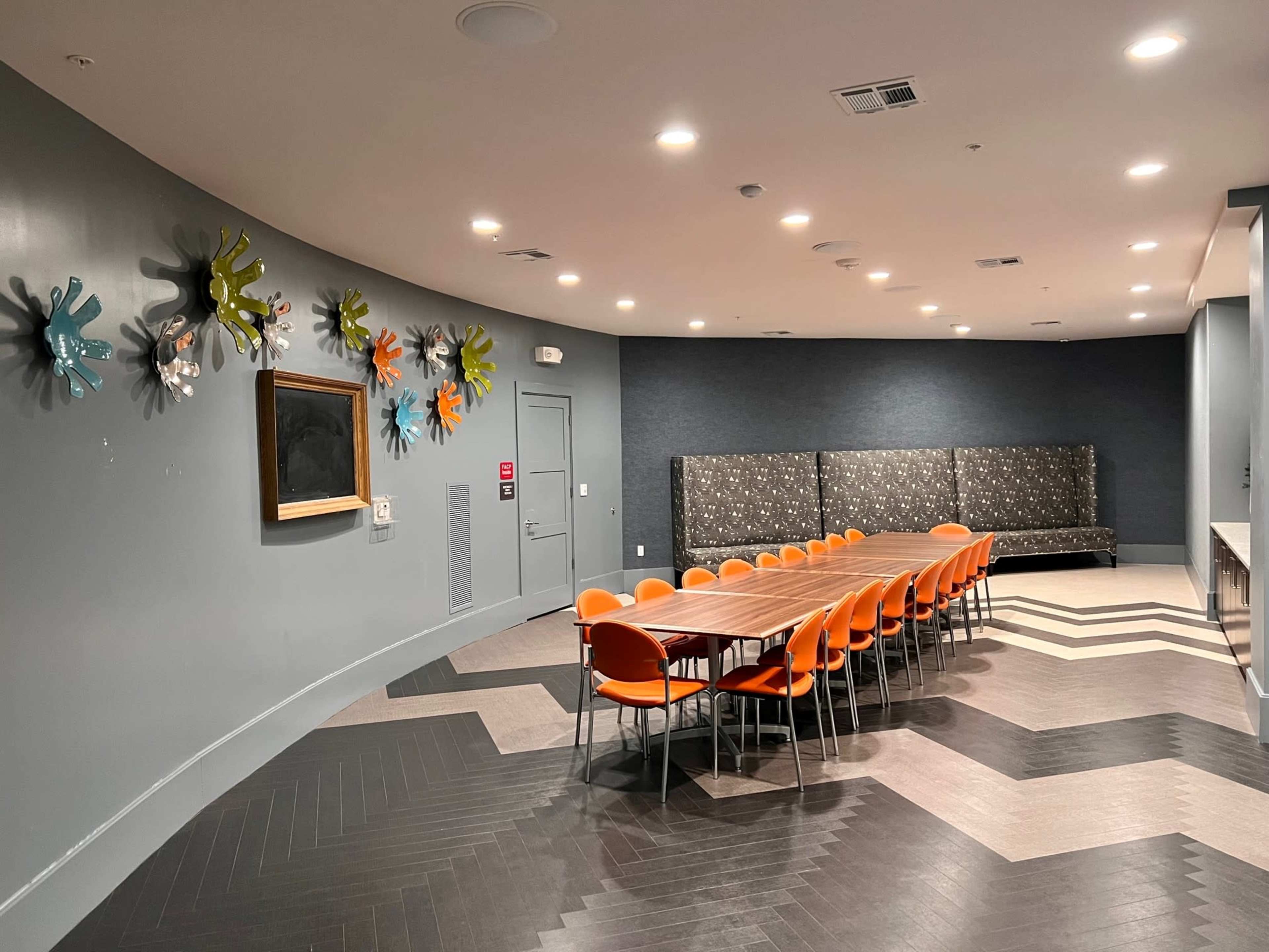 A conference room features a long wooden table surrounded by orange chairs, with colorful wall art and a seating area in the background.
