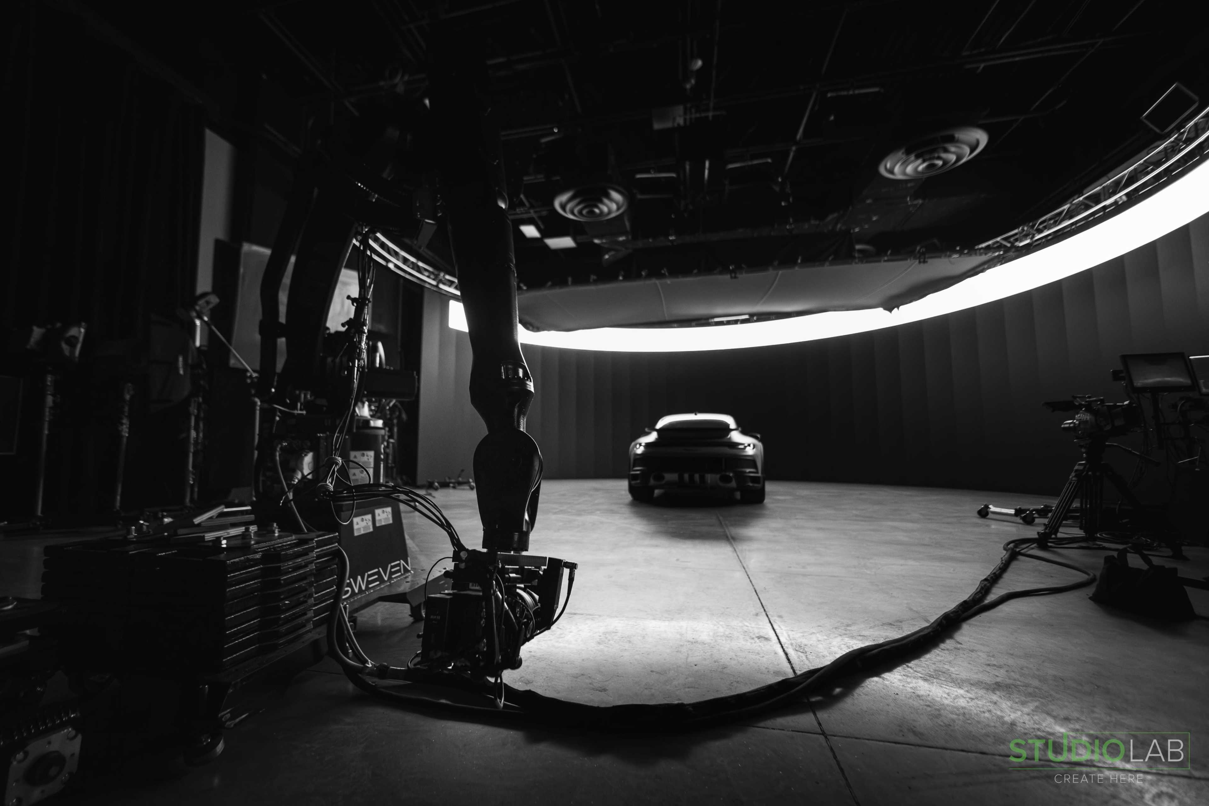 A robotic arm is positioned in front of a car in a dimly lit studio, with a curved backdrop and soft lighting illuminating the scene.