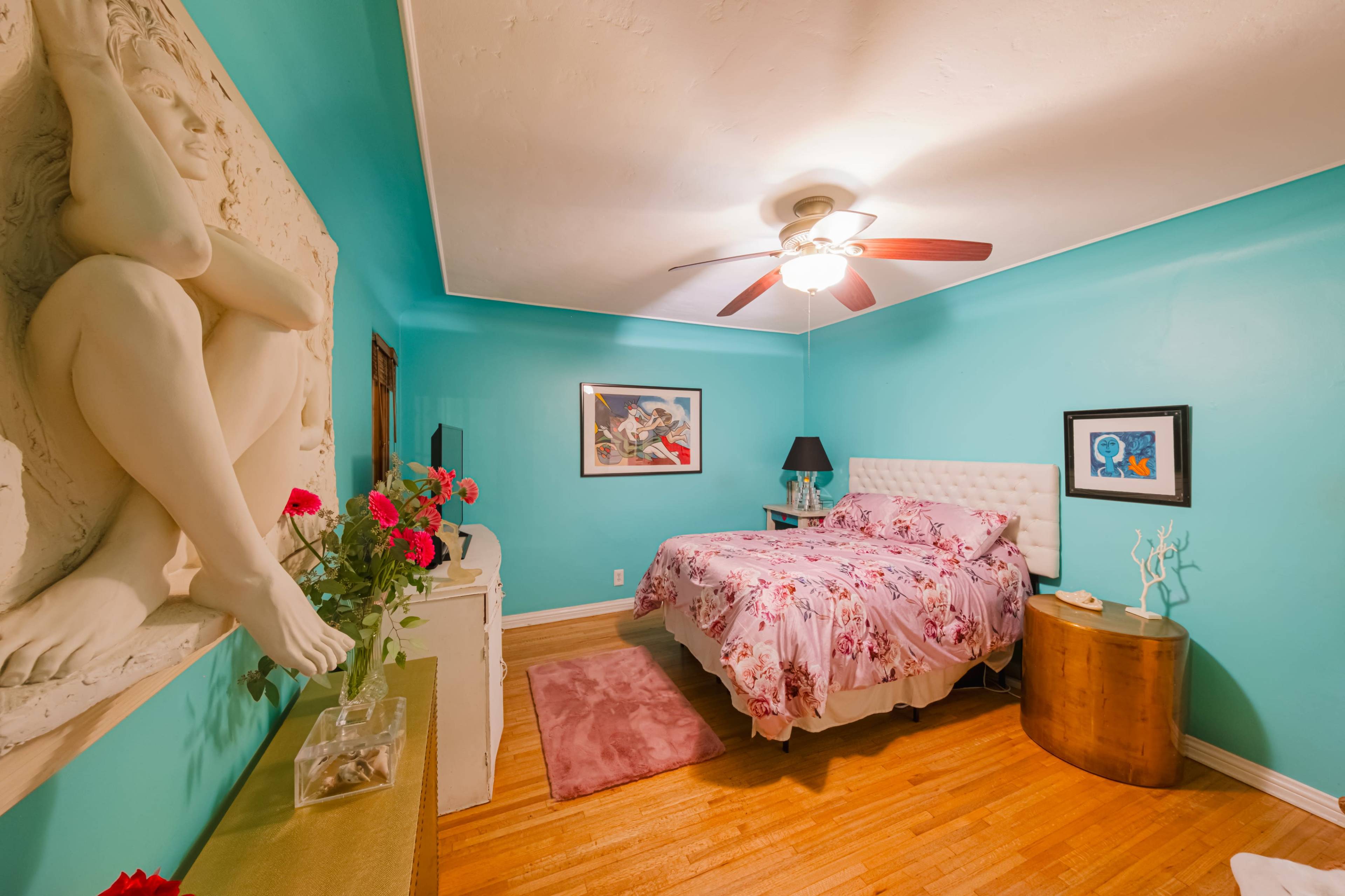 The image shows a cozy bedroom with turquoise walls, a queen-sized bed adorned with floral bedding, and a seating area featuring artistic decor.