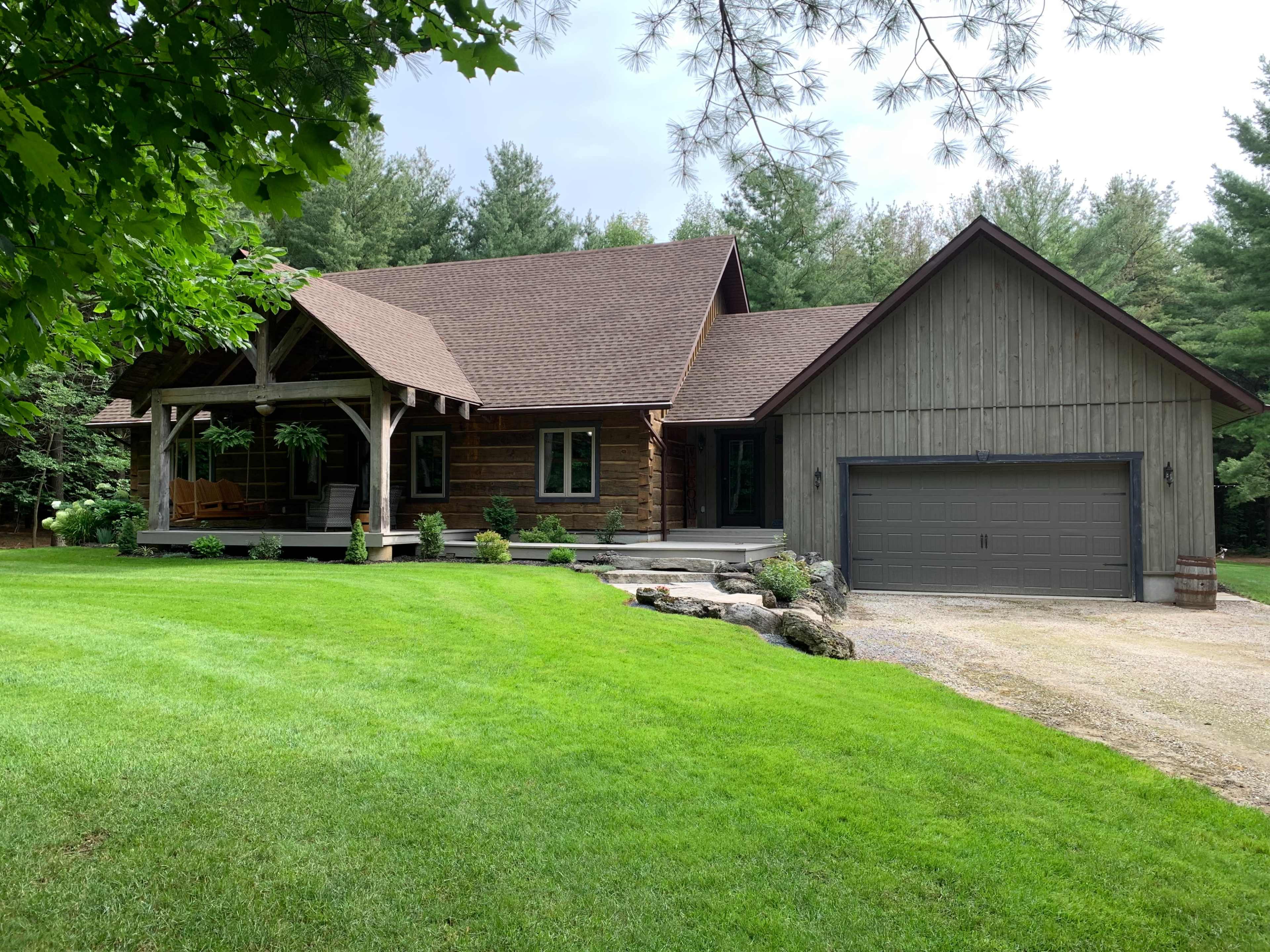 A log-style house with a gray garage sits in a grassy yard surrounded by trees.
