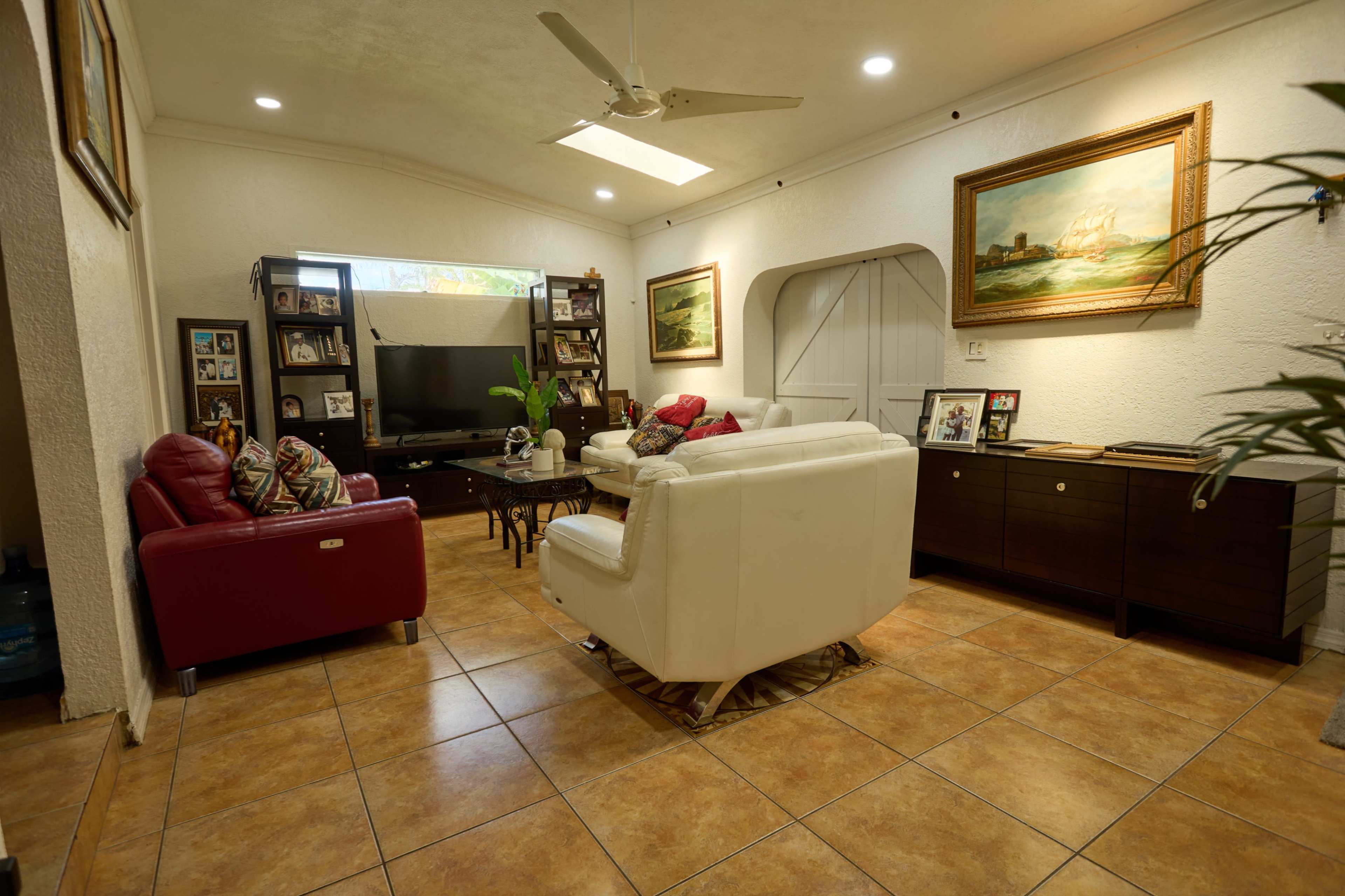 A cozy living room features a white leather sofa, a red armchair, a television set against the wall, and decorative paintings.