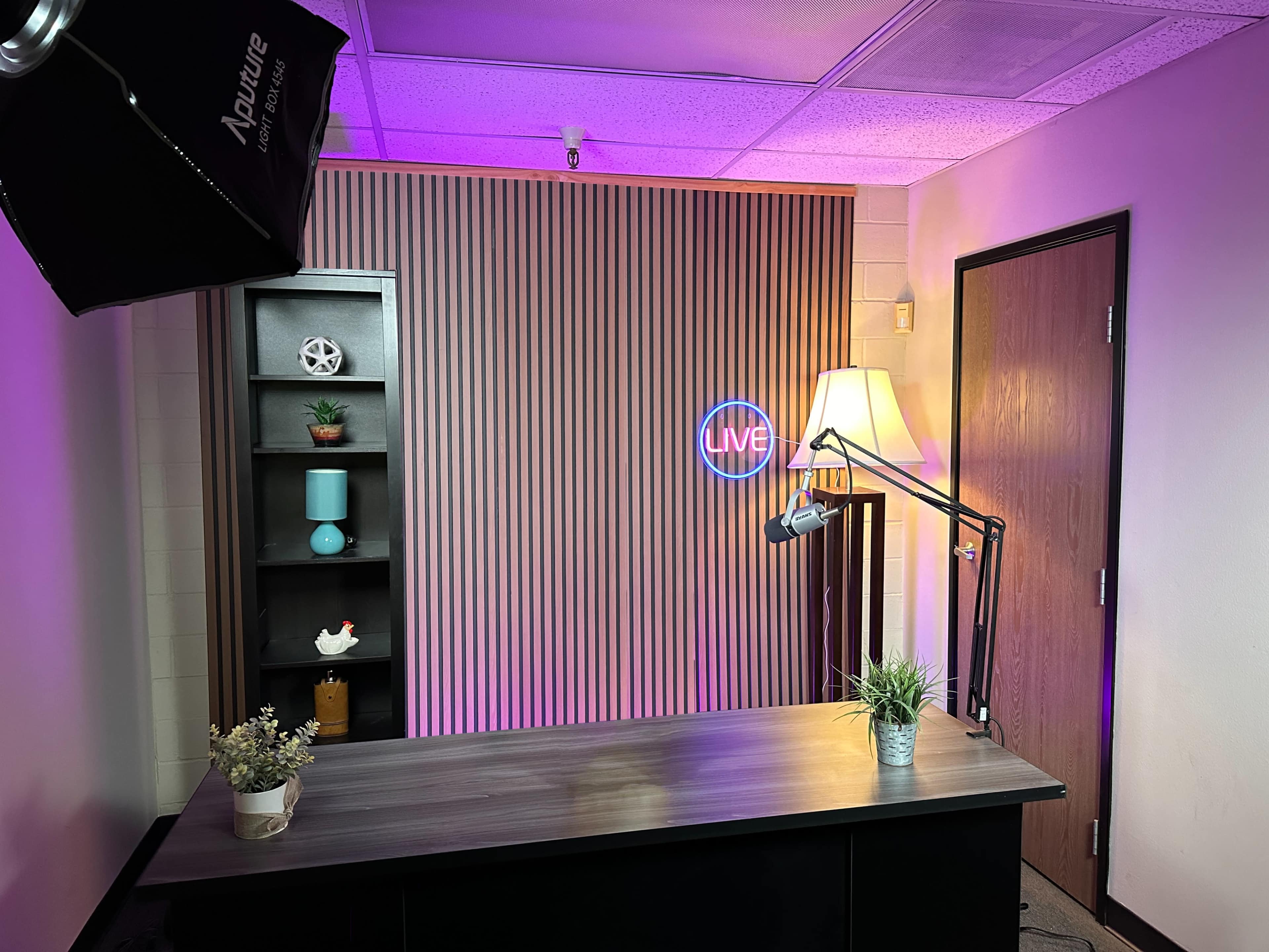A modern studio setup with a desk, a lamp, a microphone on an arm, and a decorative backdrop featuring vertical stripes and a neon "LIVE" sign.