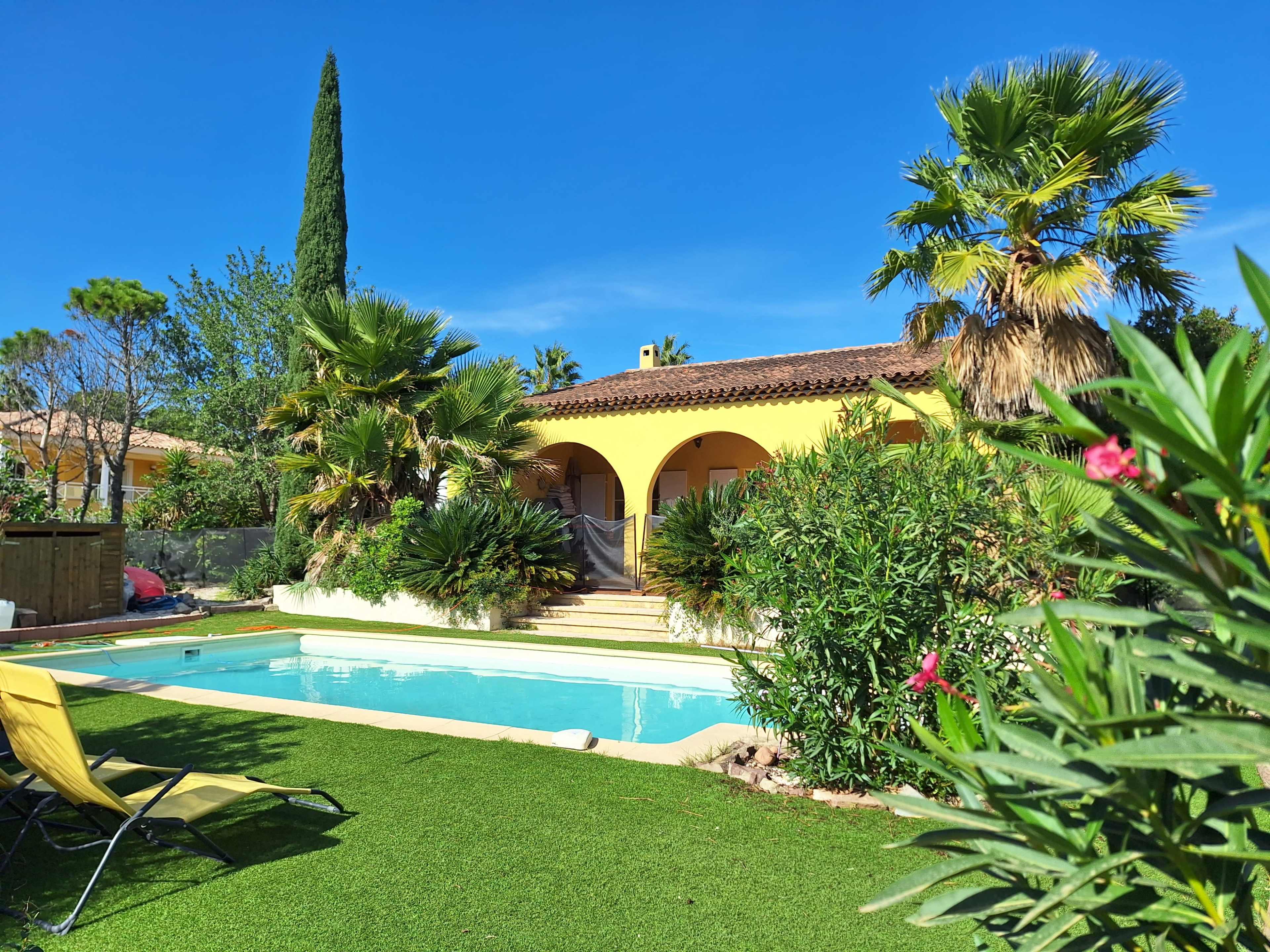 A sunny backyard features a swimming pool surrounded by tropical plants and a yellow house with arched entrances.