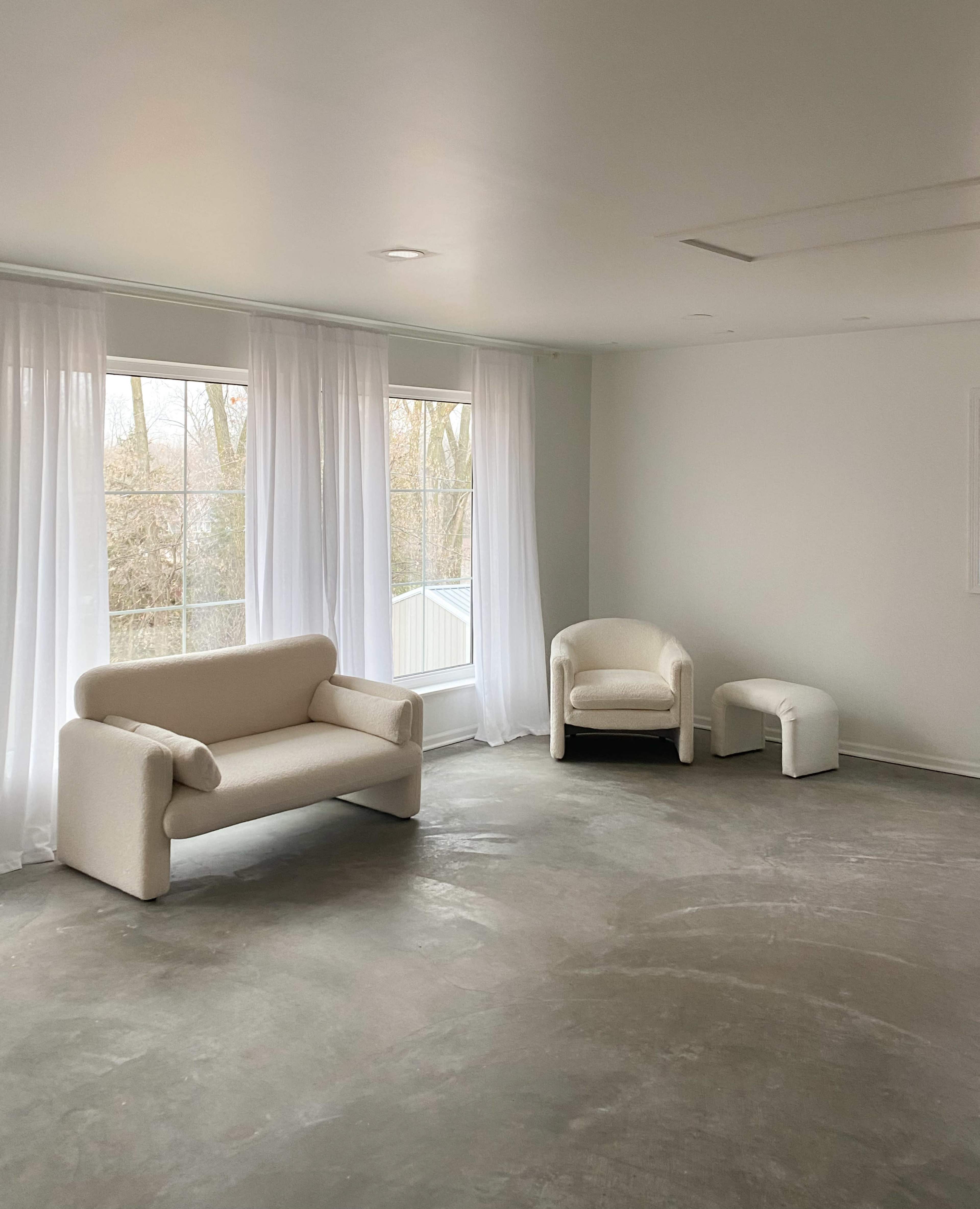 The image shows a minimalist interior with a light-colored sofa and two armchairs, accompanied by large windows covered with sheer white curtains.