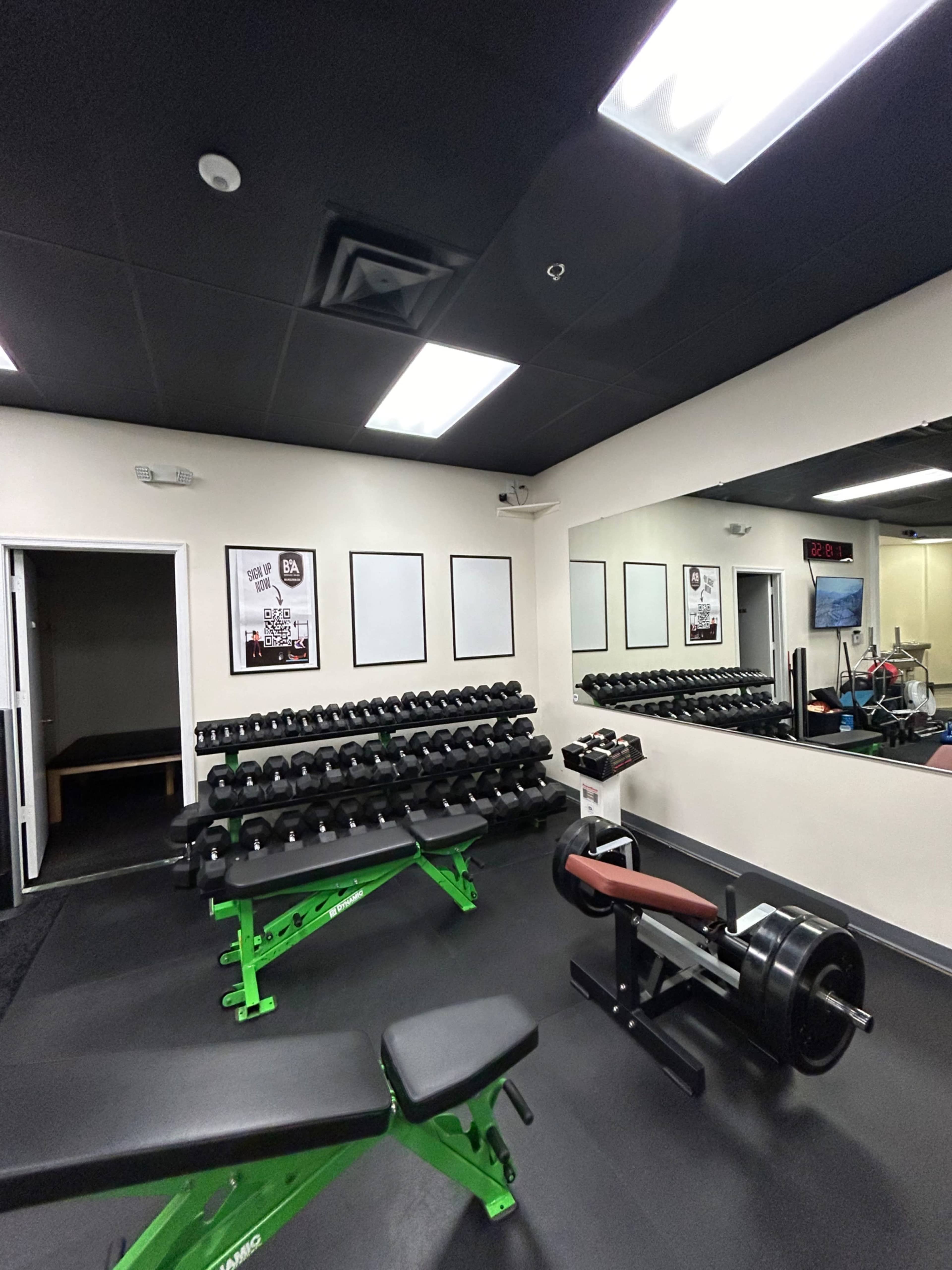 A gym interior with rows of dumbbells on a rack, a bench press, and a mirror reflecting exercise equipment.