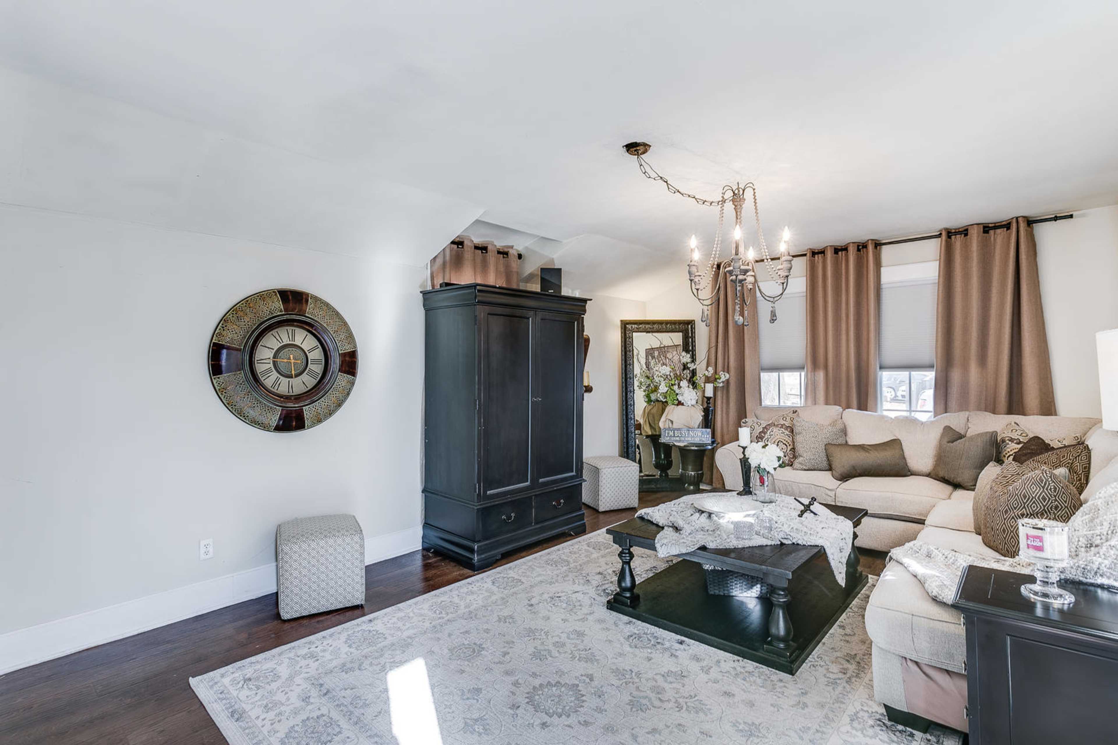 A living room featuring a light-colored sofa, a dark wooden coffee table, a large decorative wall clock, and a chandelier hanging from the ceiling.