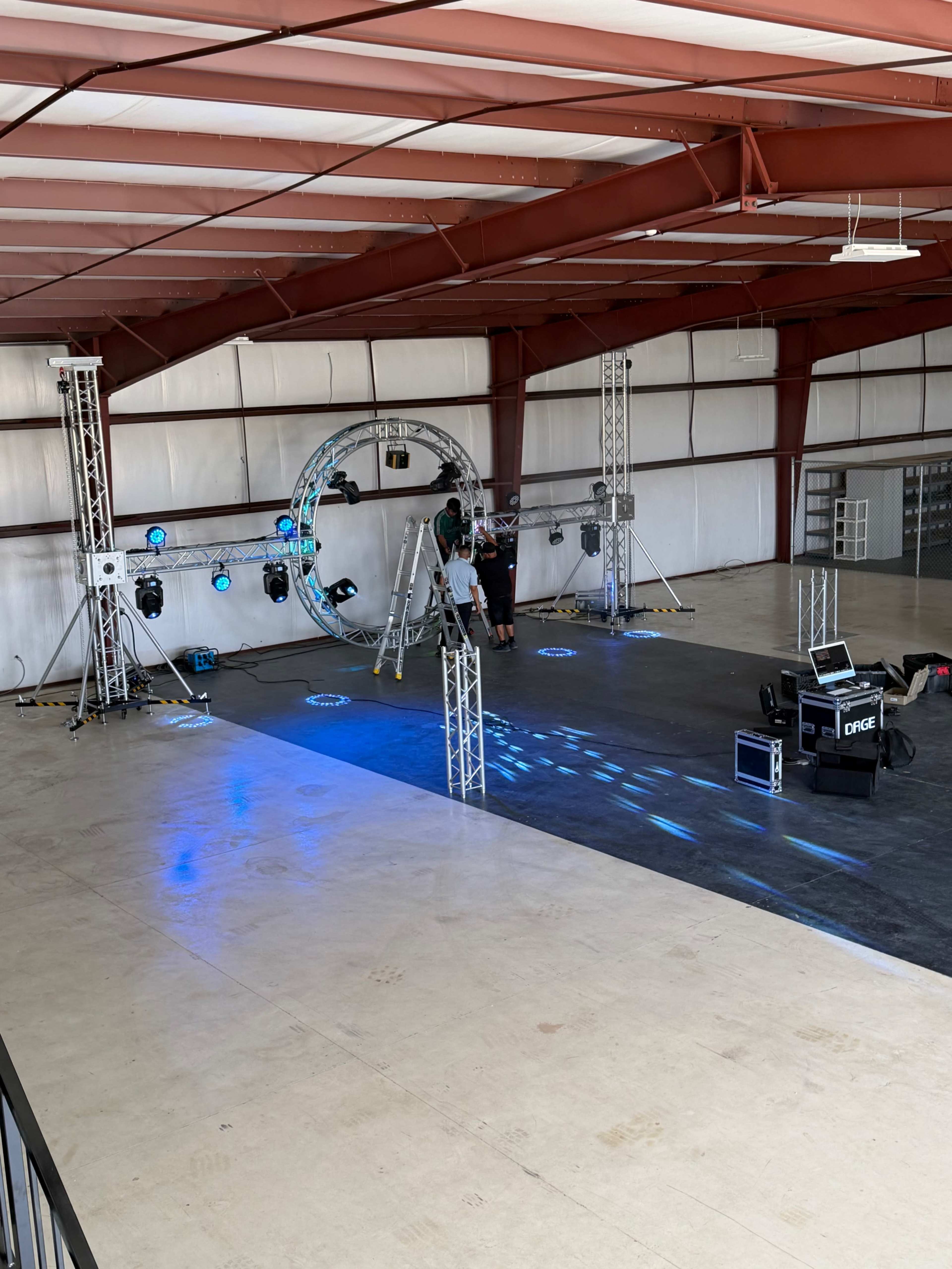A partially set up event space features a stage with lighting fixtures and audio equipment in a spacious metal building.