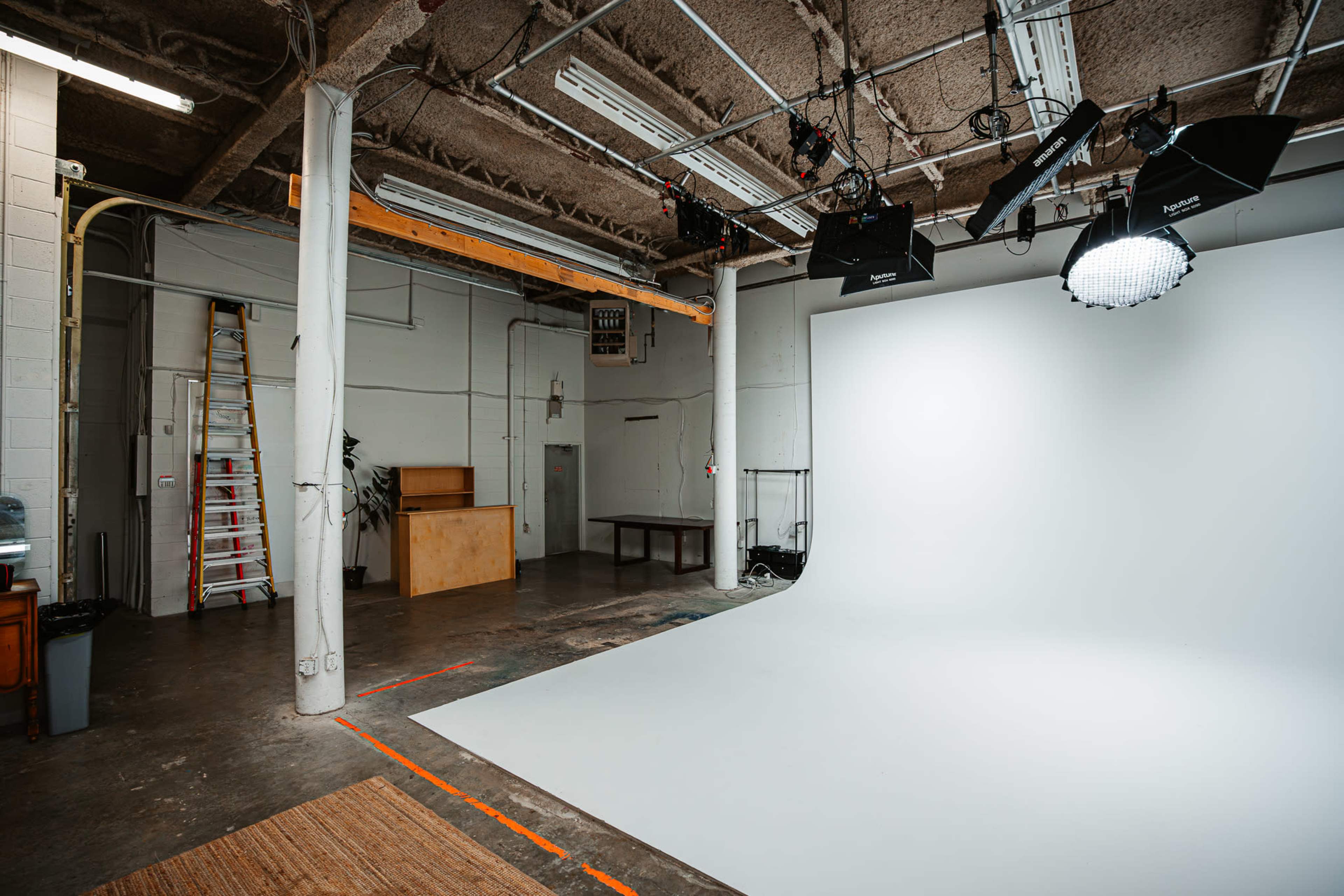 The image shows a spacious photography studio with a white backdrop, a ladder, a wooden table, and studio lighting equipment.