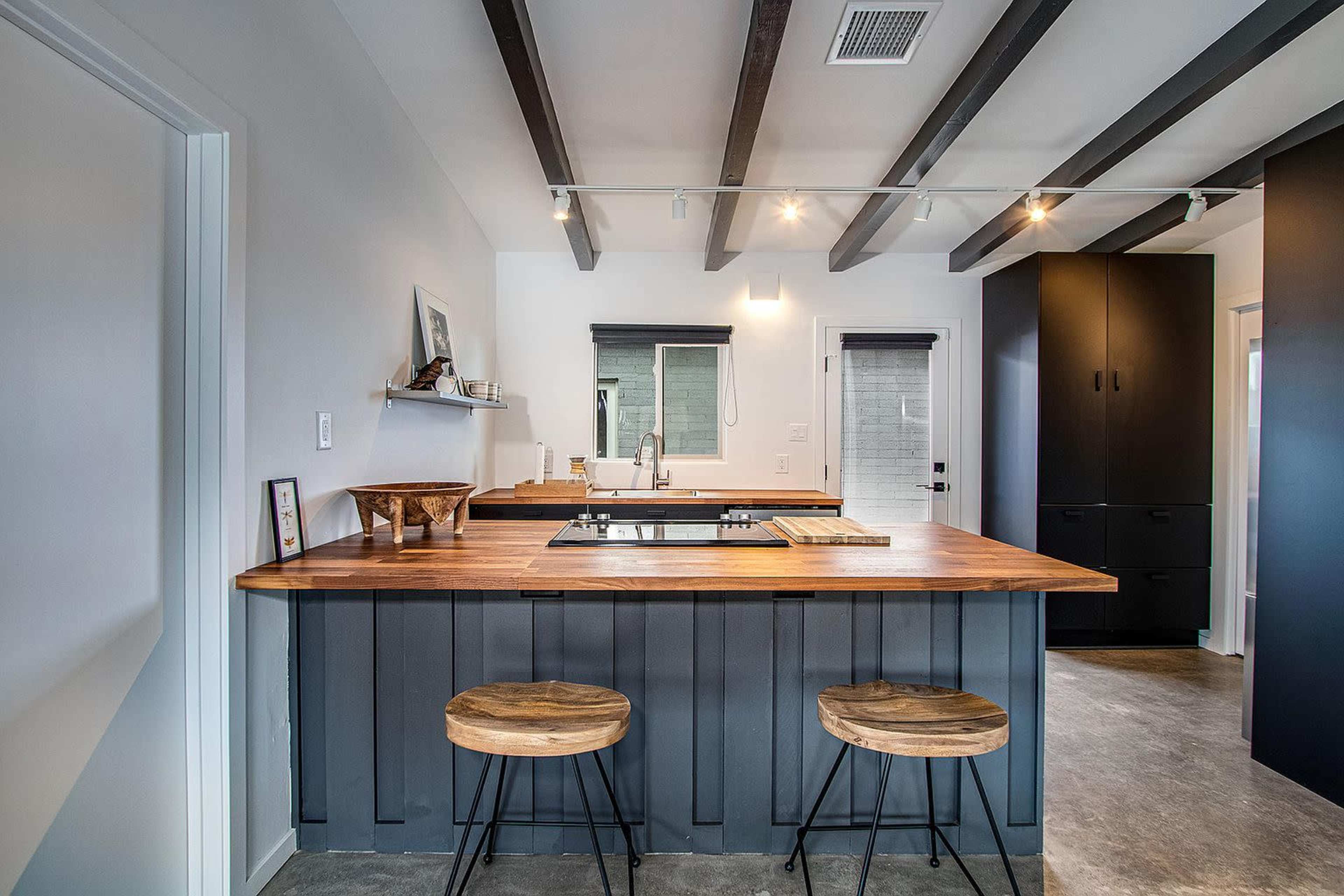 A modern kitchen with a wooden island, two stools, and contrasting dark cabinetry against light walls and ceiling beams.