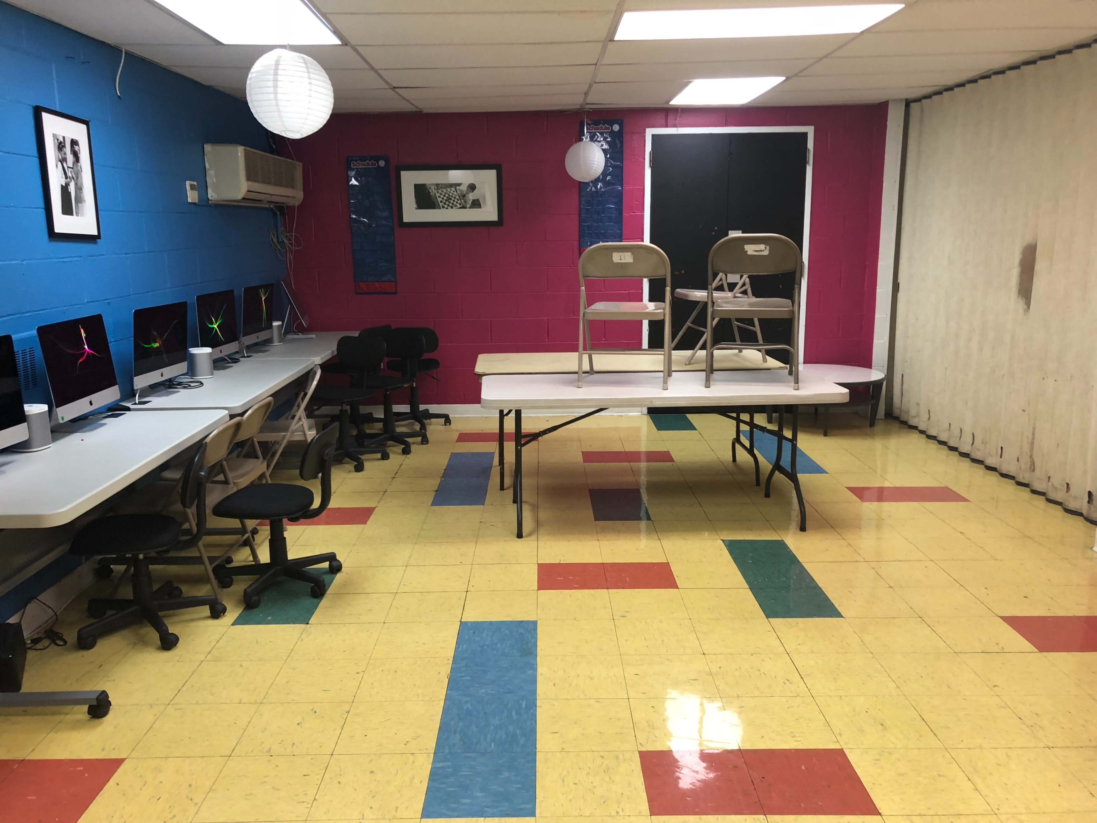 A computer room with rows of desks, chairs, and a table displaying stacked chairs, set against a brightly colored wall.