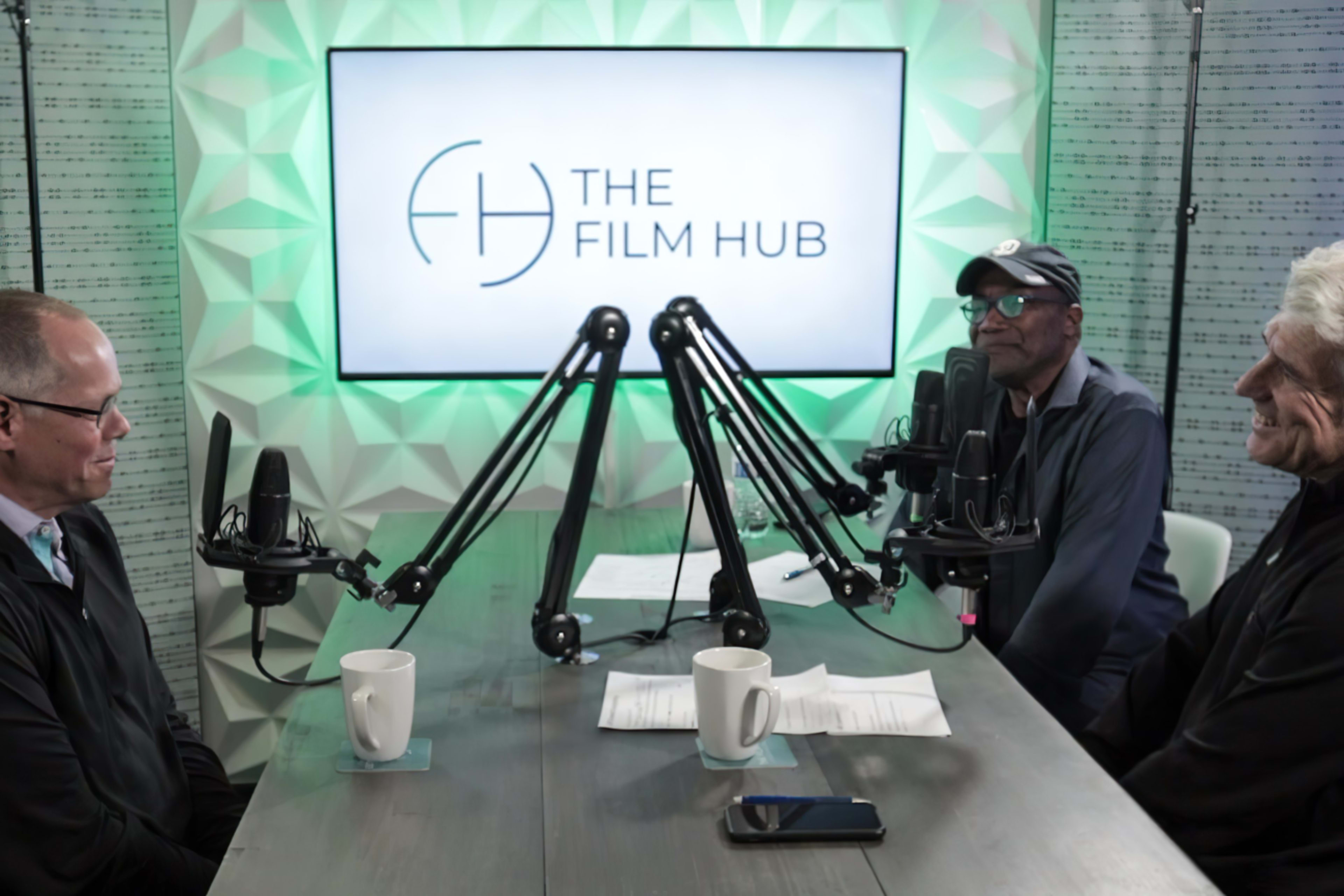 Three individuals are seated at a table with microphones, discussing in front of a screen displaying "THE FILM HUB."