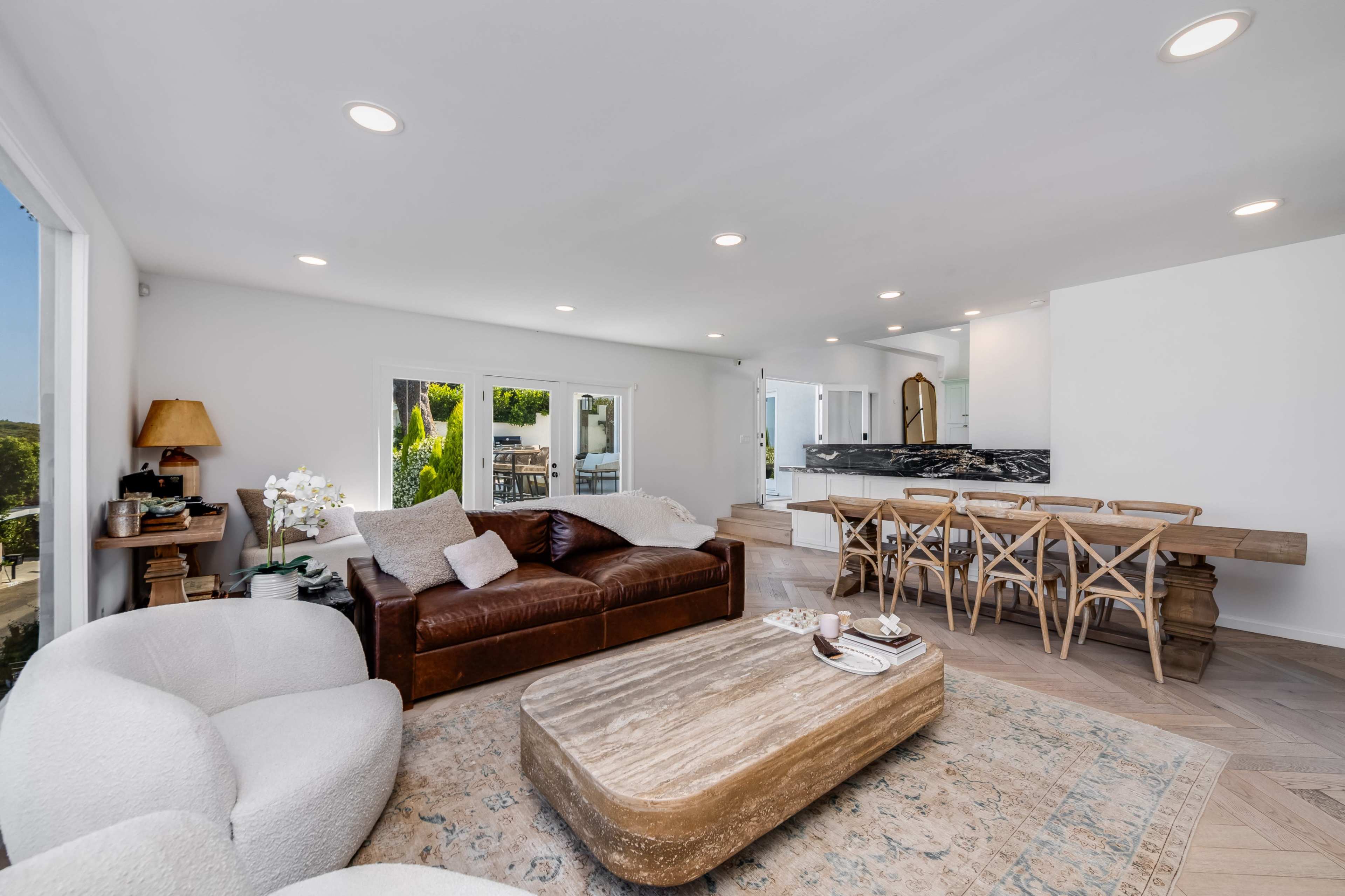 The image shows a modern living room with a brown leather sofa, a light-colored armchair, a wooden coffee table, and a dining area with a light wood table and chairs.