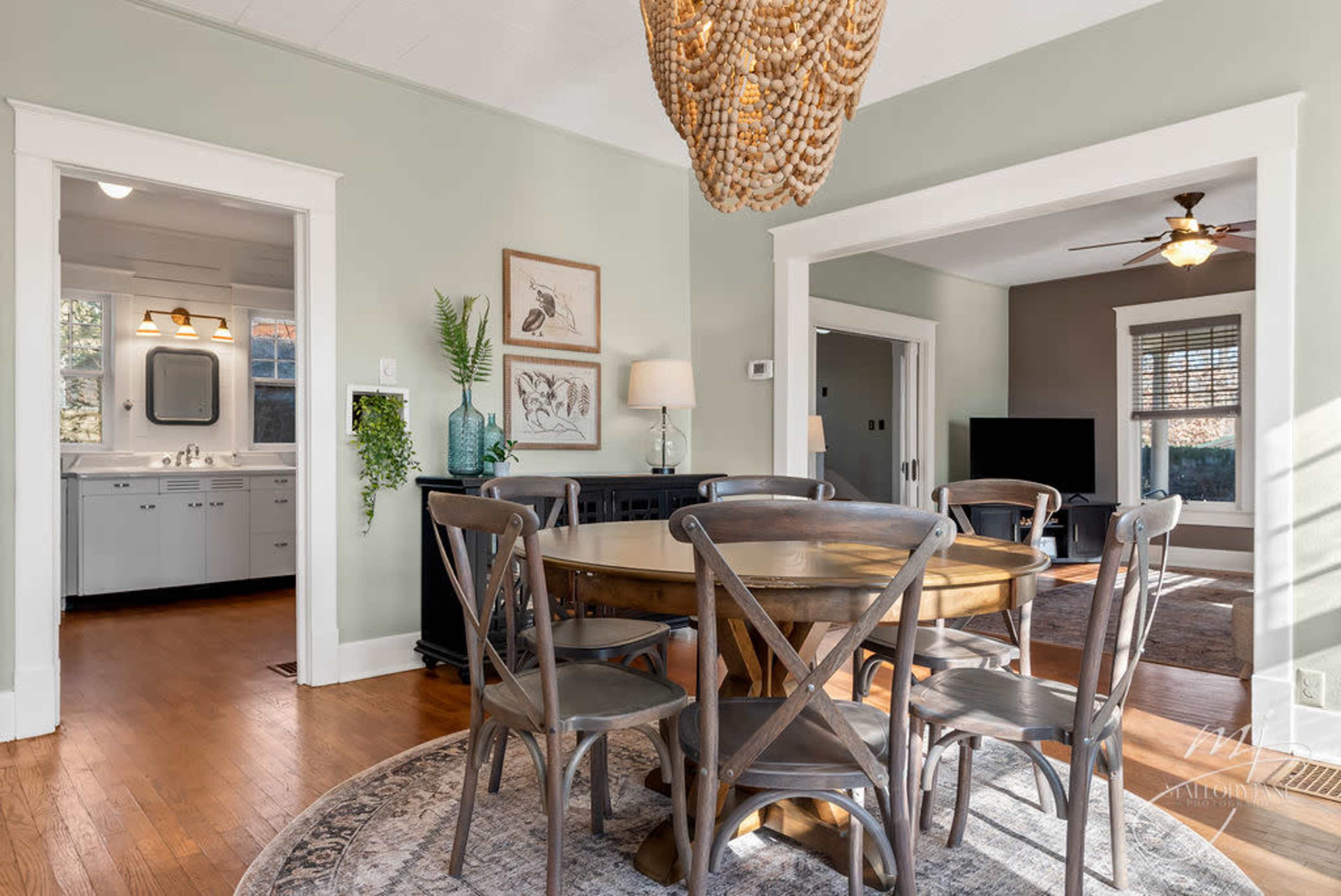 A dining room features a wooden table surrounded by gray chairs, with a large light fixture hanging overhead and doorways leading to adjacent rooms.