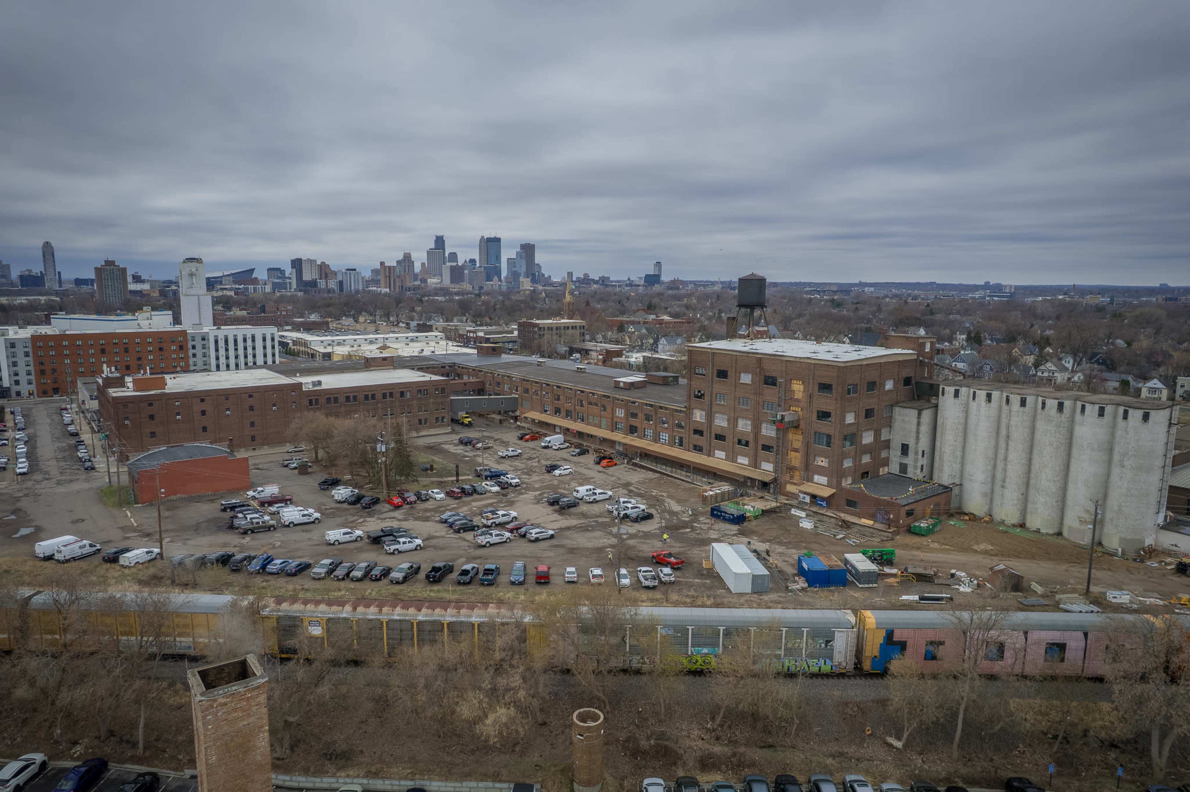 The image shows an aerial view of a large, abandoned industrial area with several buildings and parked vehicles, set against a skyline of a city in the background.