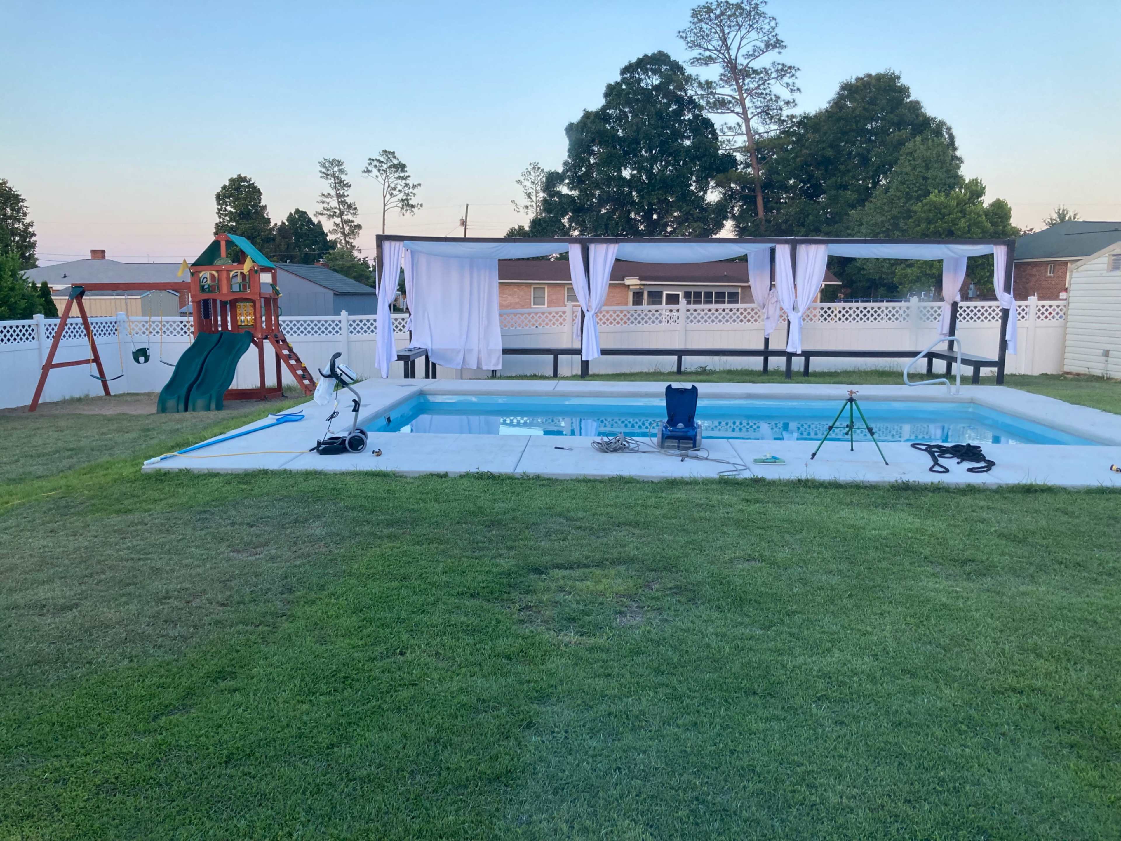 A backyard with a swimming pool, a covered seating area, a playground structure, and green grass.