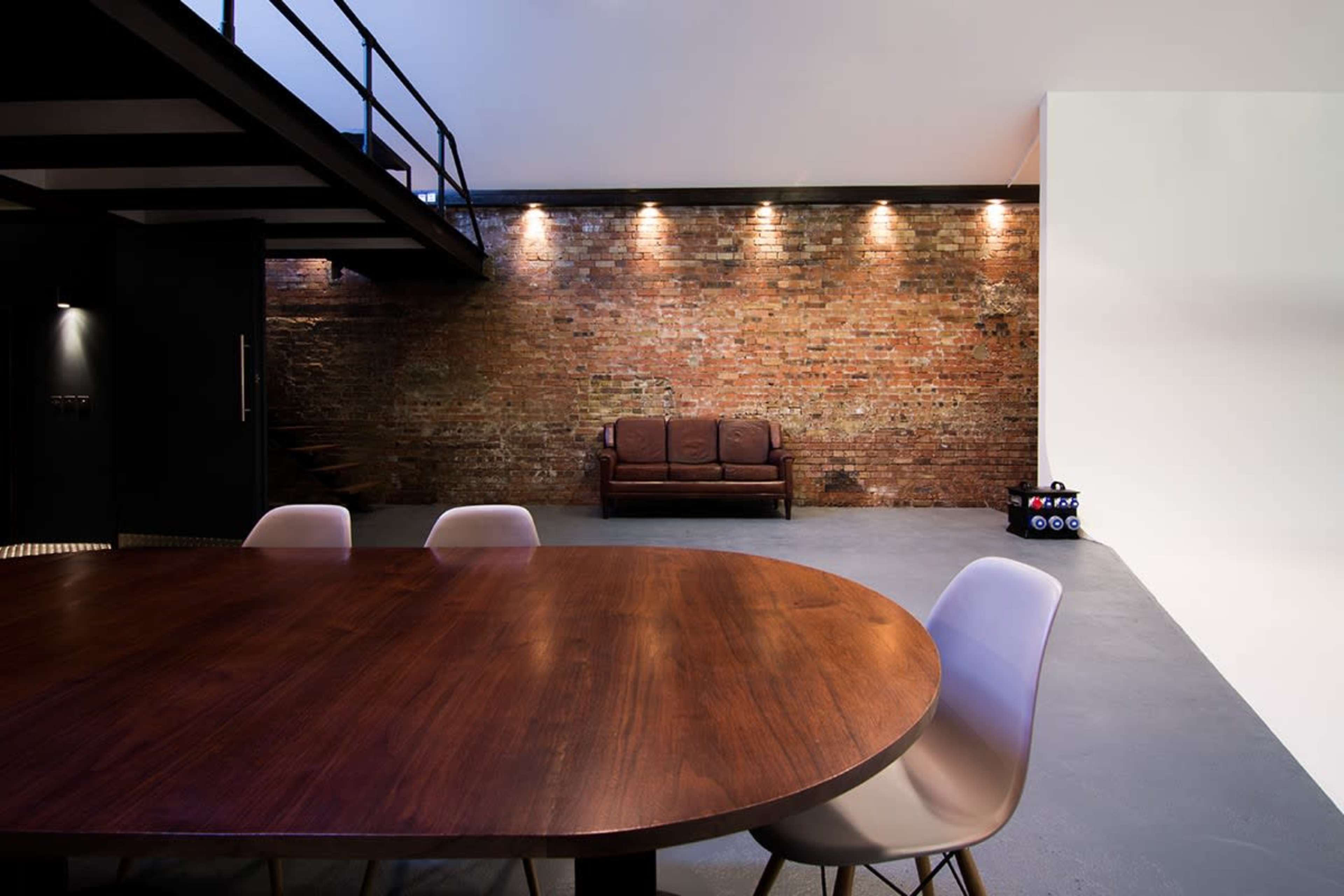 The image features a spacious interior with a brown leather sofa against a brick wall, a large round wooden dining table, and sleek modern chairs.
