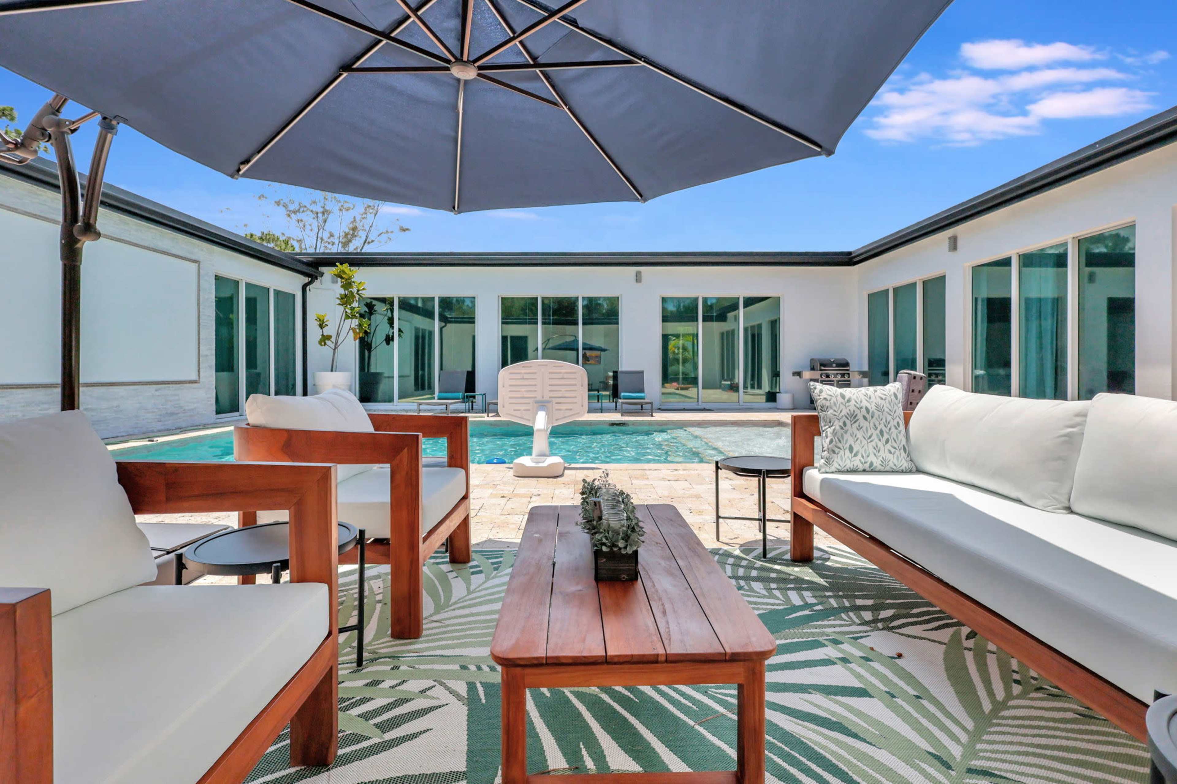 A backyard seating area with wooden furniture and a large umbrella beside a swimming pool surrounded by glass-walled rooms.