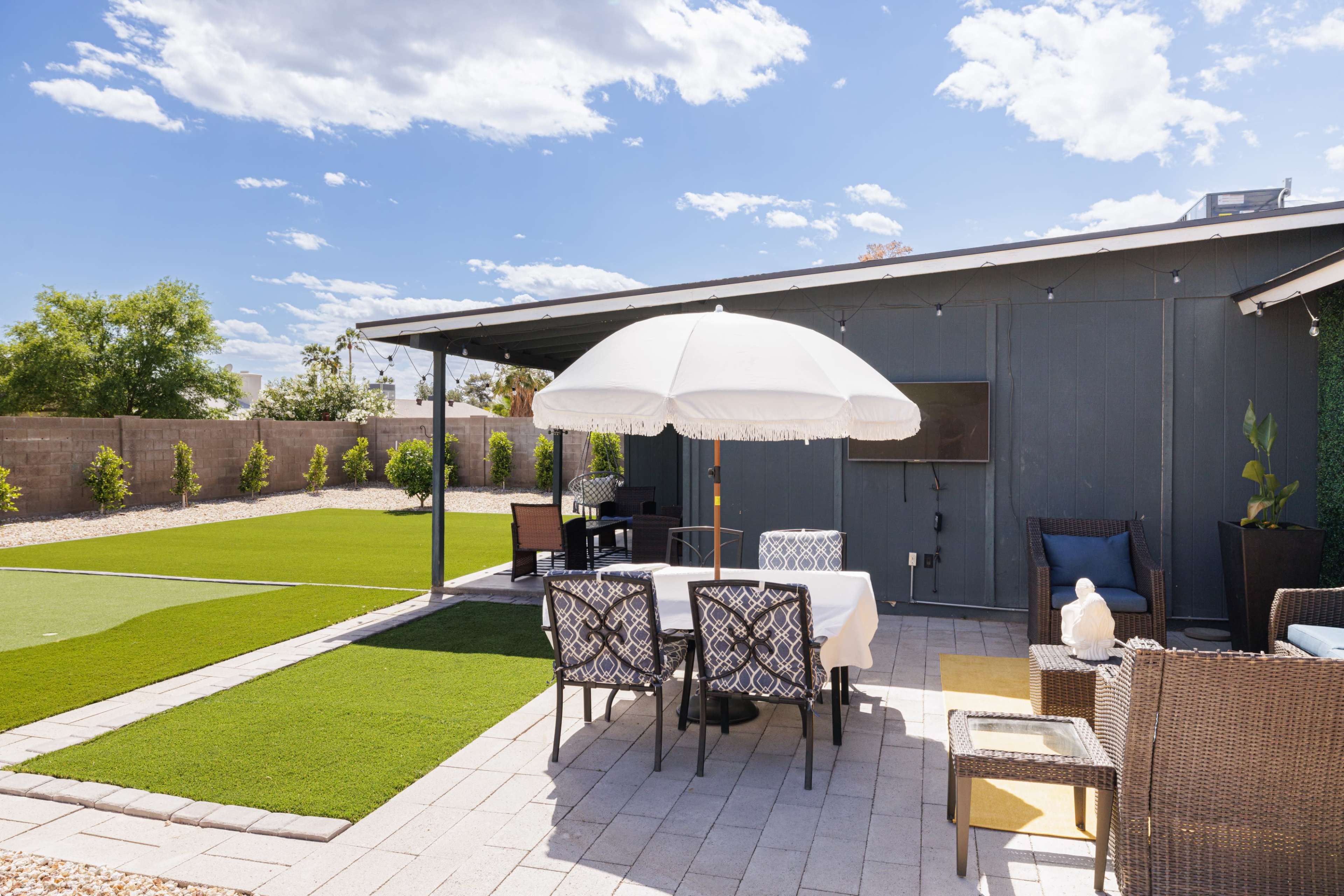 A patio area features a white umbrella over a dining table surrounded by six chairs, with artificial turf and landscaped greenery in the background.