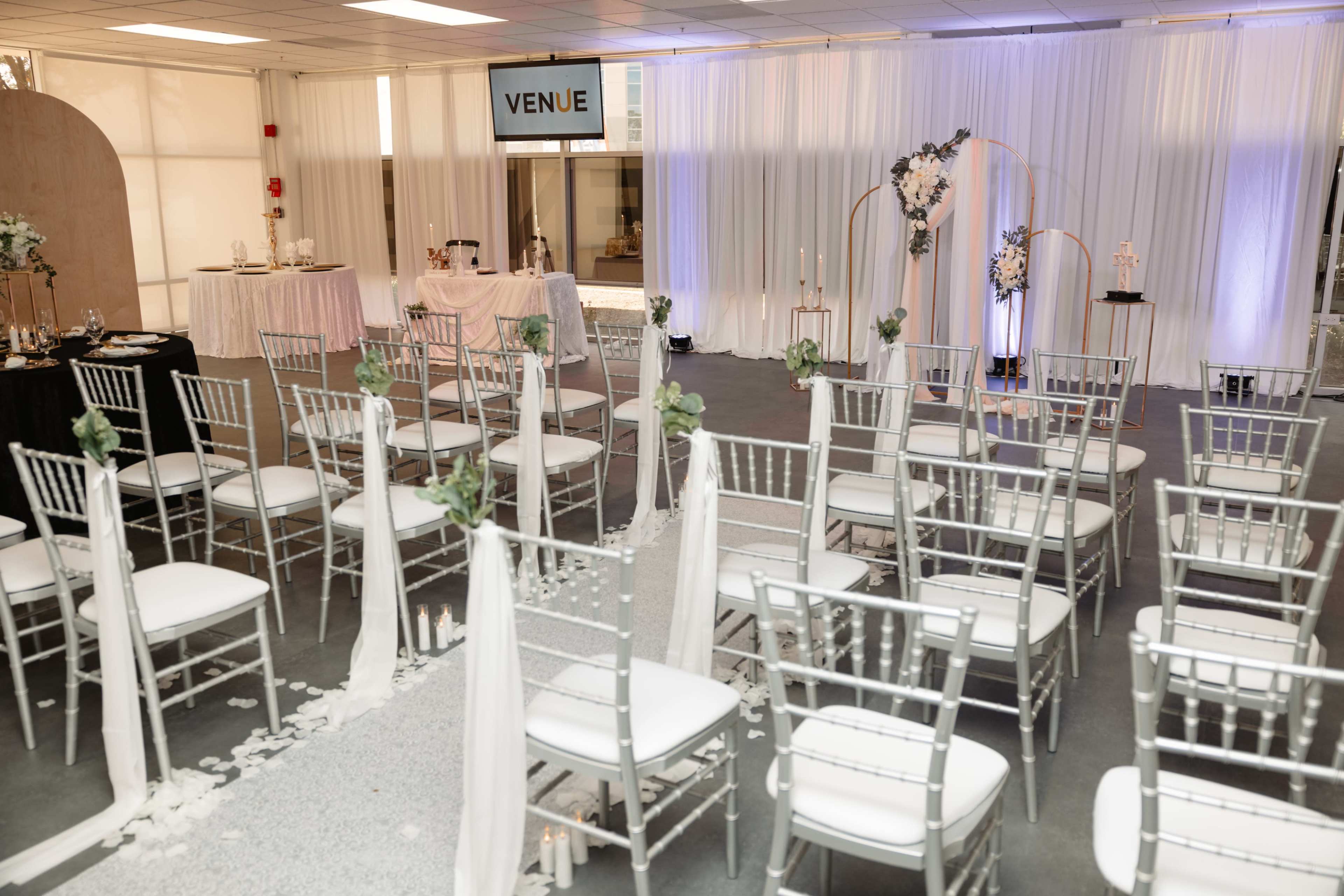 The image shows a wedding ceremony setup in a modern venue with silver chairs arranged in rows, a decorated altar area, and a table adorned with floral arrangements.