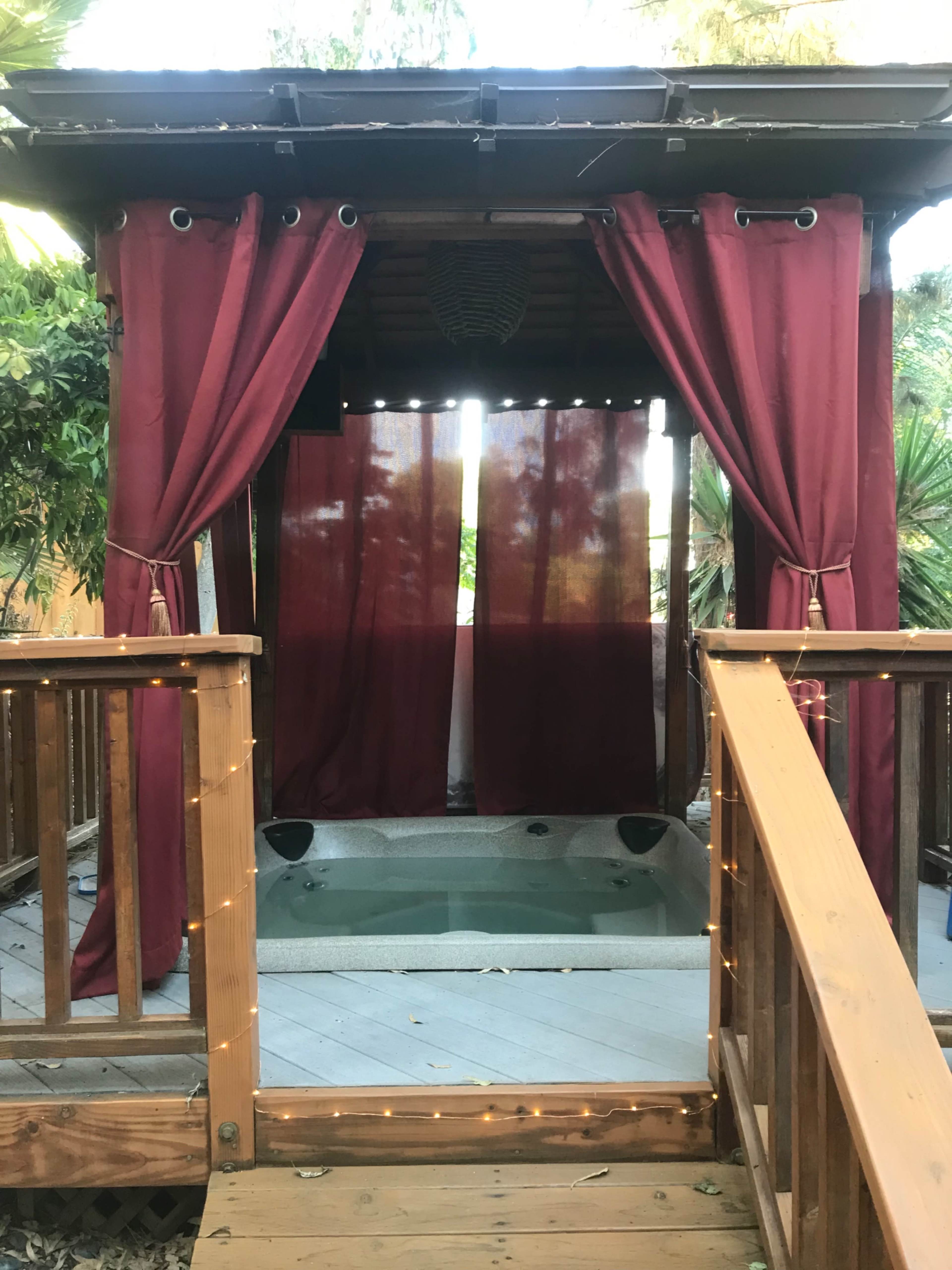 A wooden deck leads to a hot tub enclosed by sheer curtains and draped fabric, with surrounding greenery visible.