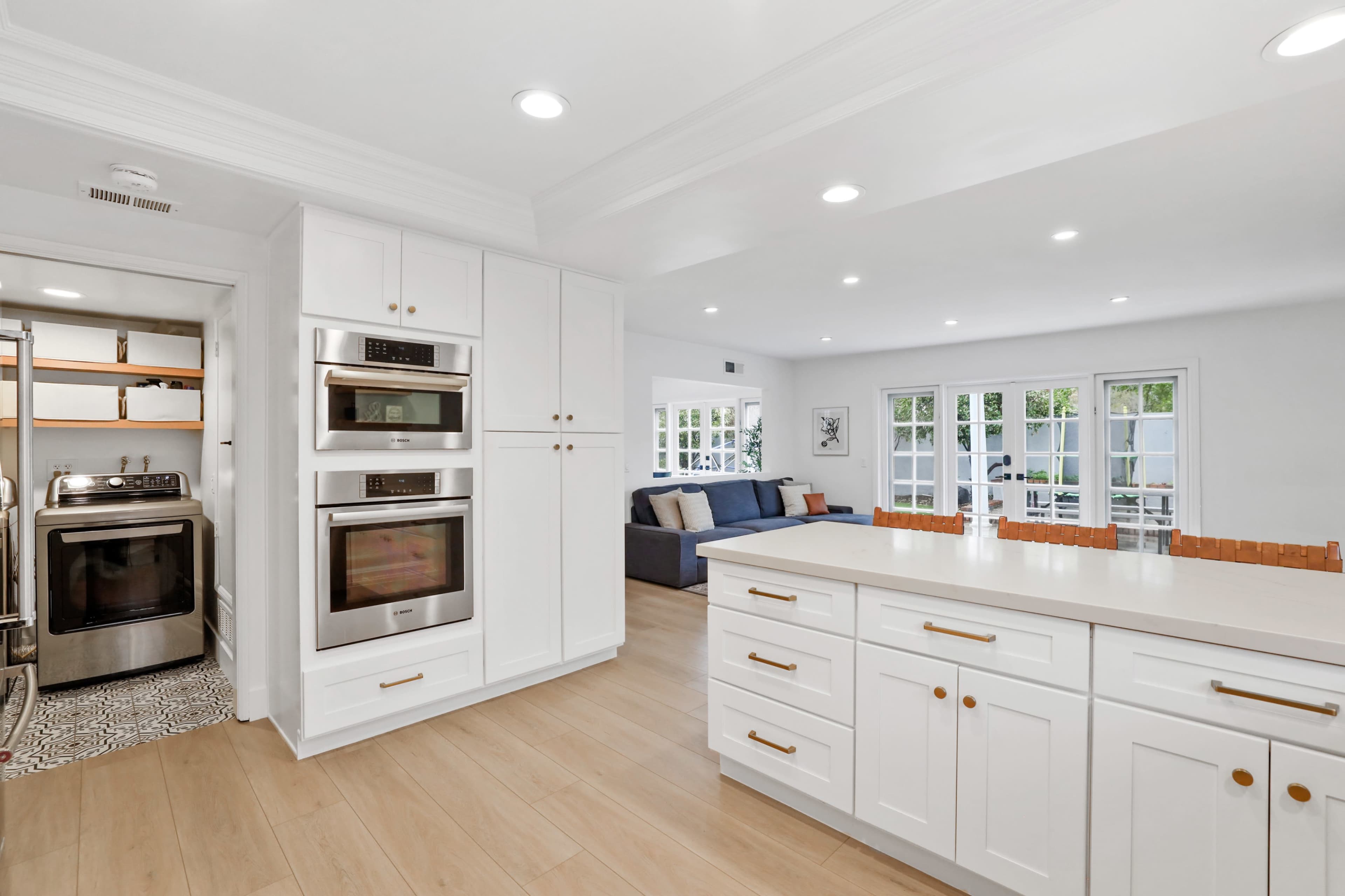 A modern kitchen with white cabinetry, stainless steel double ovens, and a view of a connected living area featuring large windows.