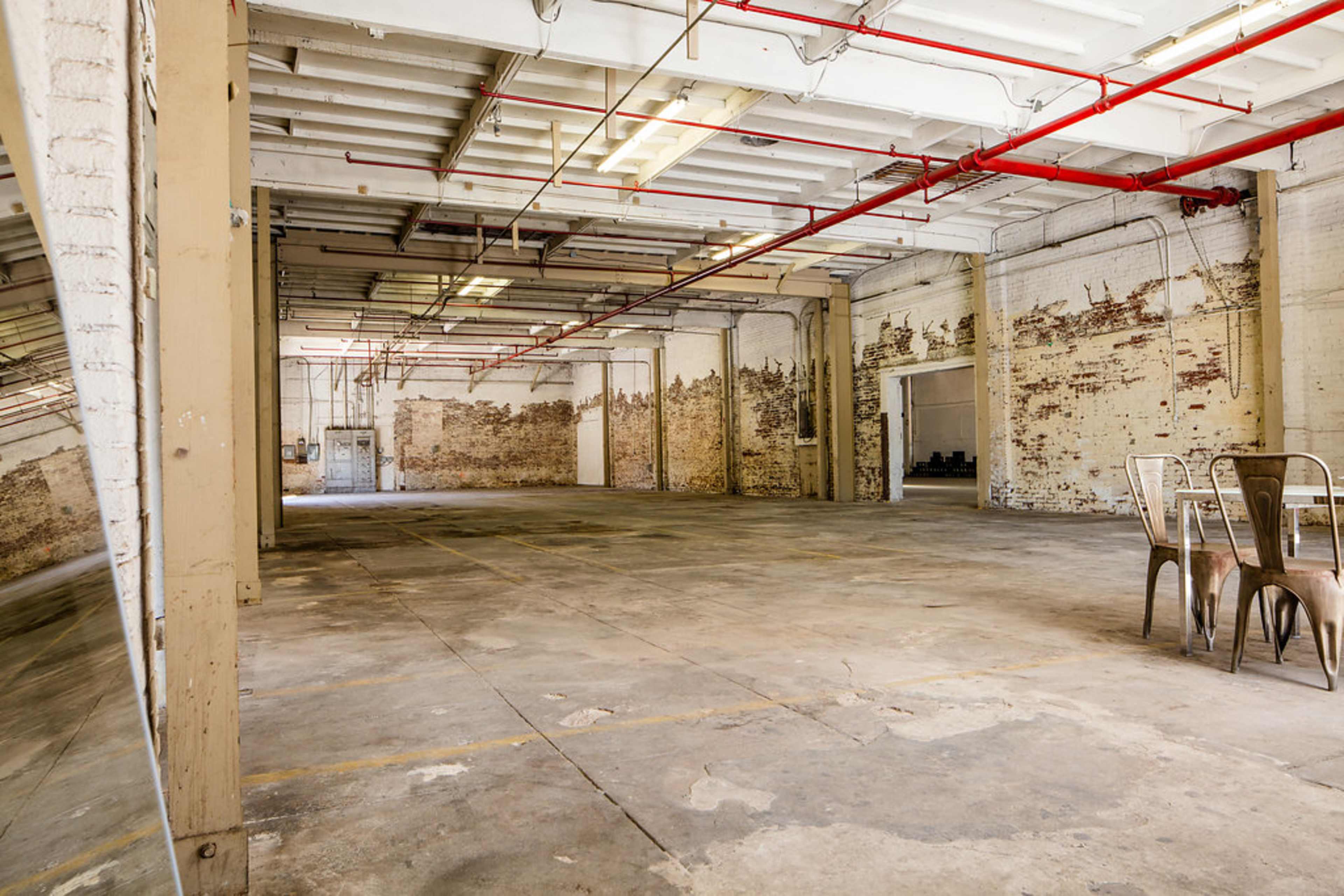 An expansive, vacant industrial space features exposed brick walls, concrete flooring, and overhead pipes within a well-lit environment.
