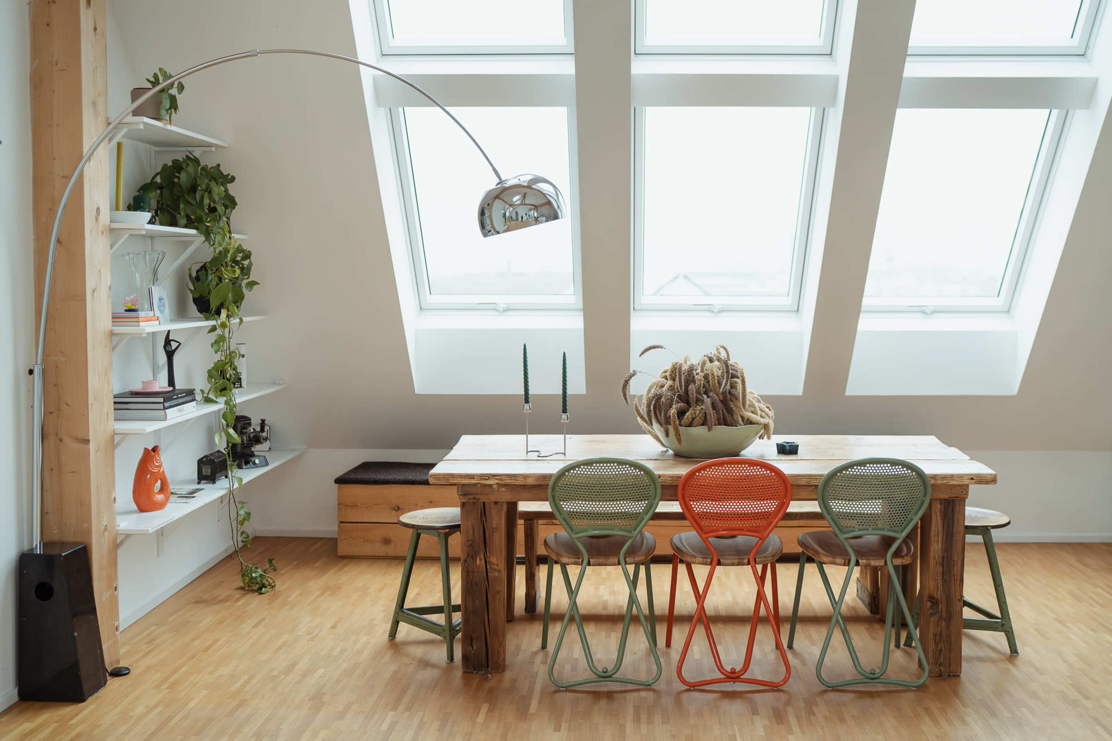 The image shows a modern dining area featuring a large wooden table surrounded by colorful chairs, a minimalist shelf with plants, and large windows that provide ample natural light.