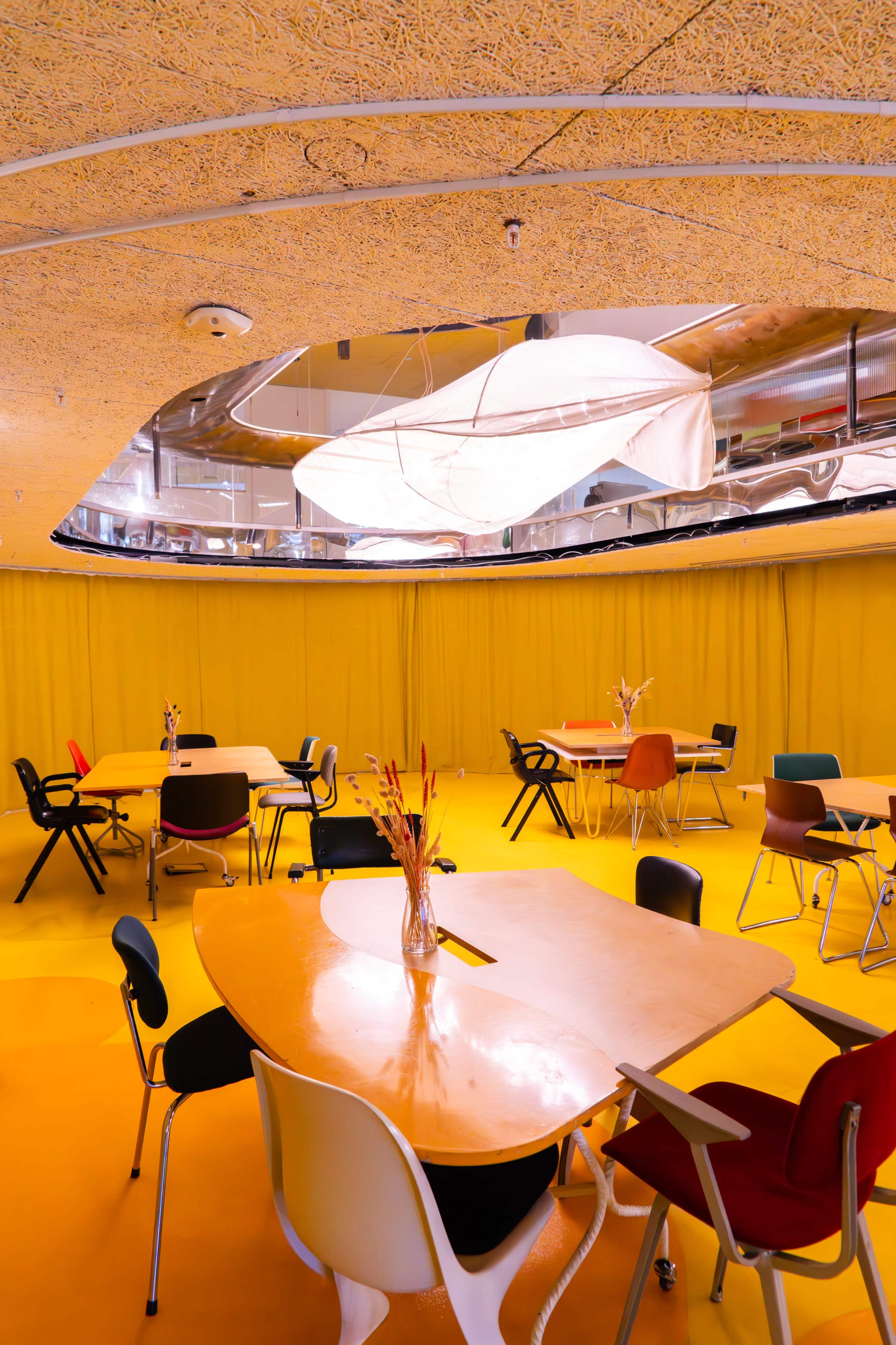 The interior of a modern café features round tables with a mix of colorful chairs against a bright yellow floor and walls, illuminated by overhead lighting.