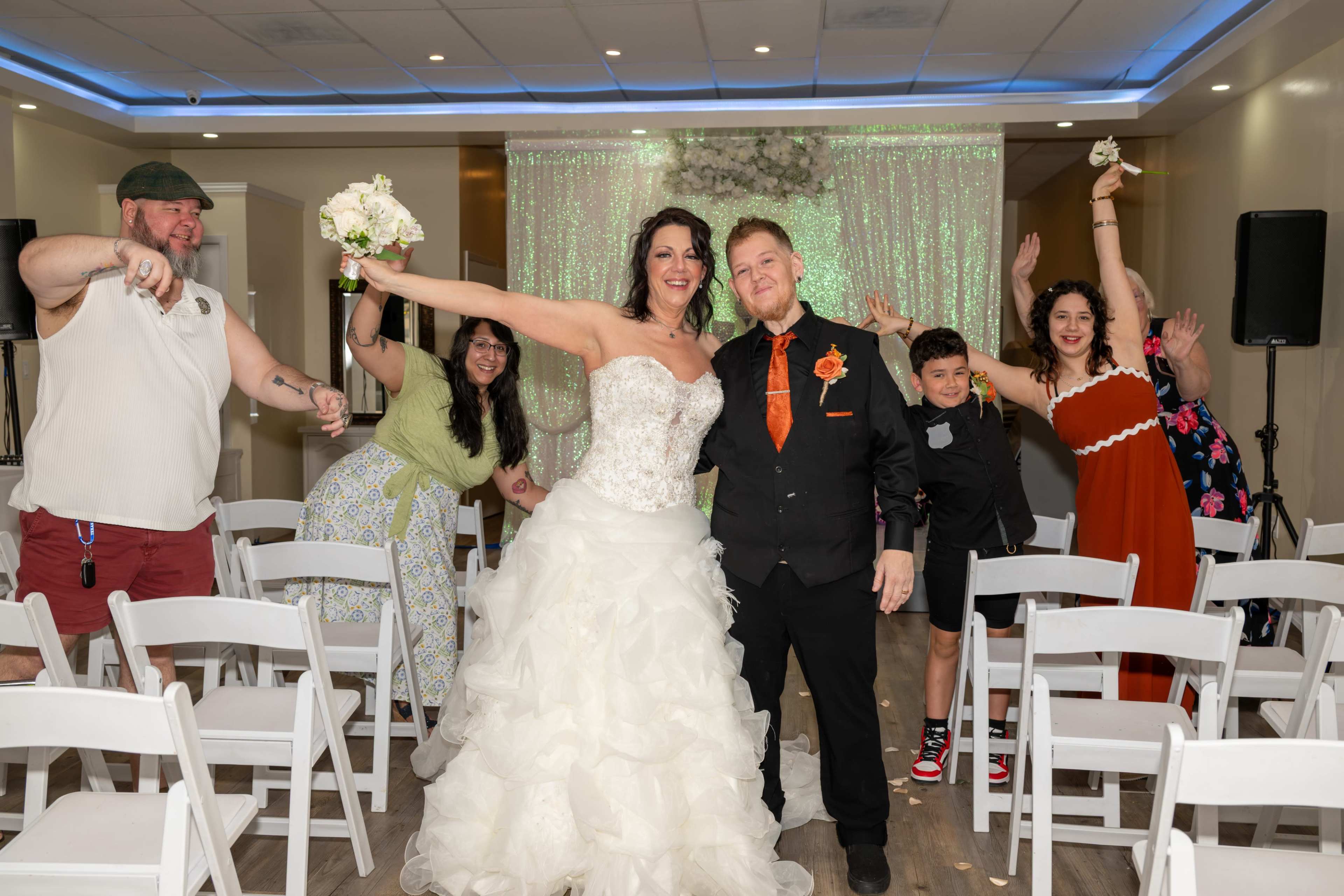 A bride and groom pose for a photo with their guests in a decorated indoor space set up for a wedding ceremony.