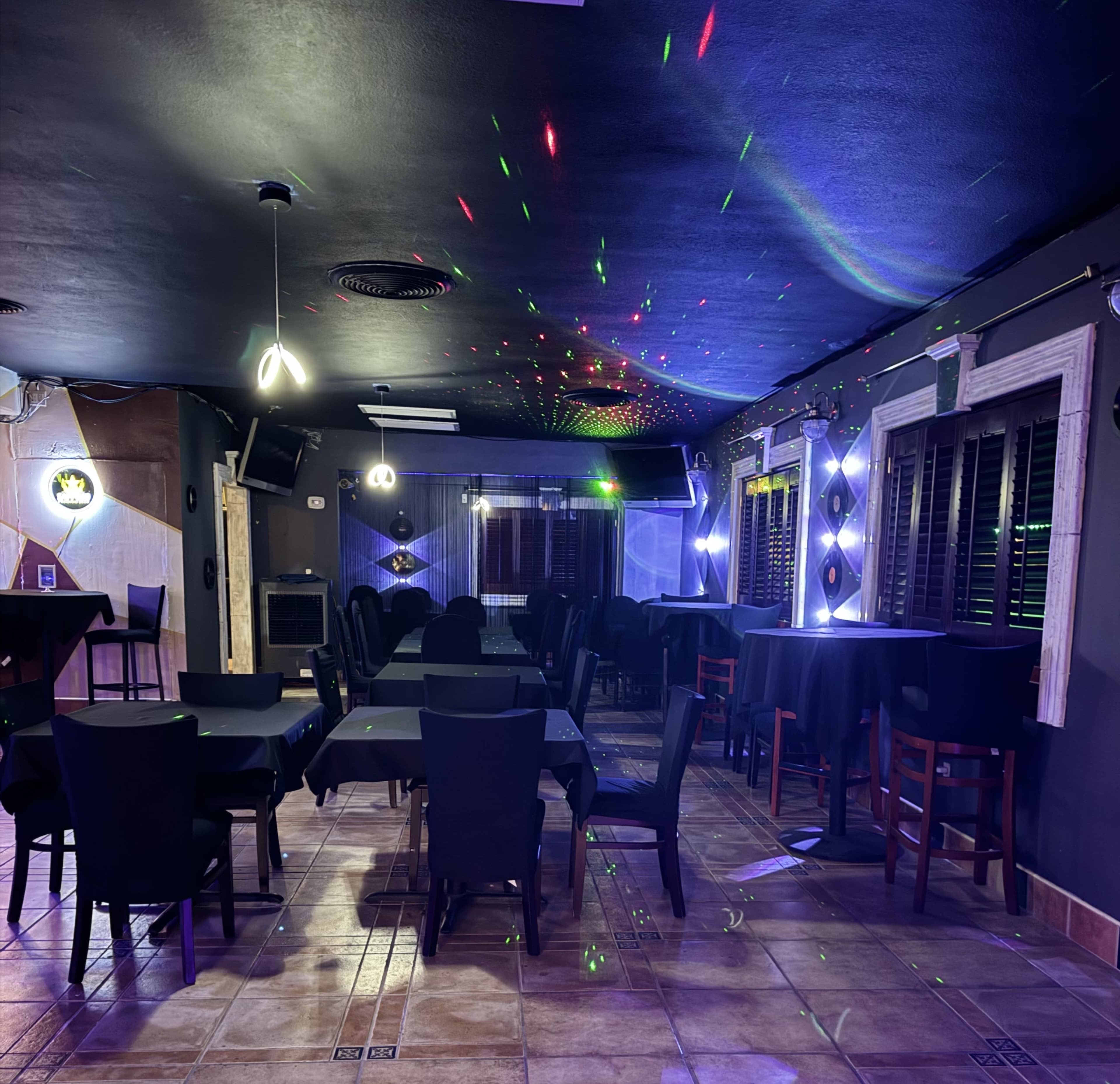 The interior of a dimly lit restaurant features black tables and chairs, with colorful lights reflecting on the walls and ceiling.