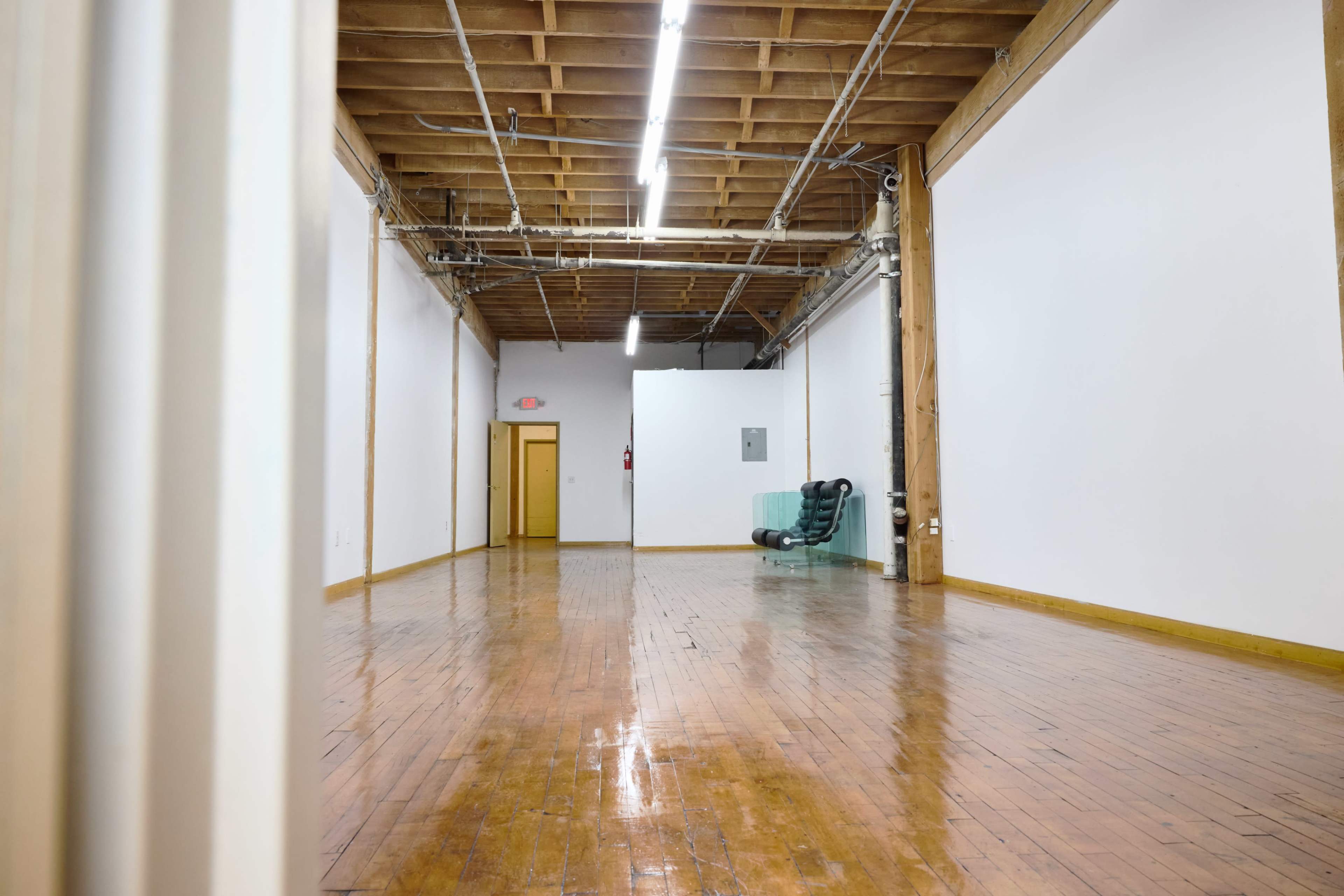 The image shows a spacious, empty room with wooden flooring, high ceilings, and exposed beams, featuring a green chair against one wall and a doorway leading to another area.