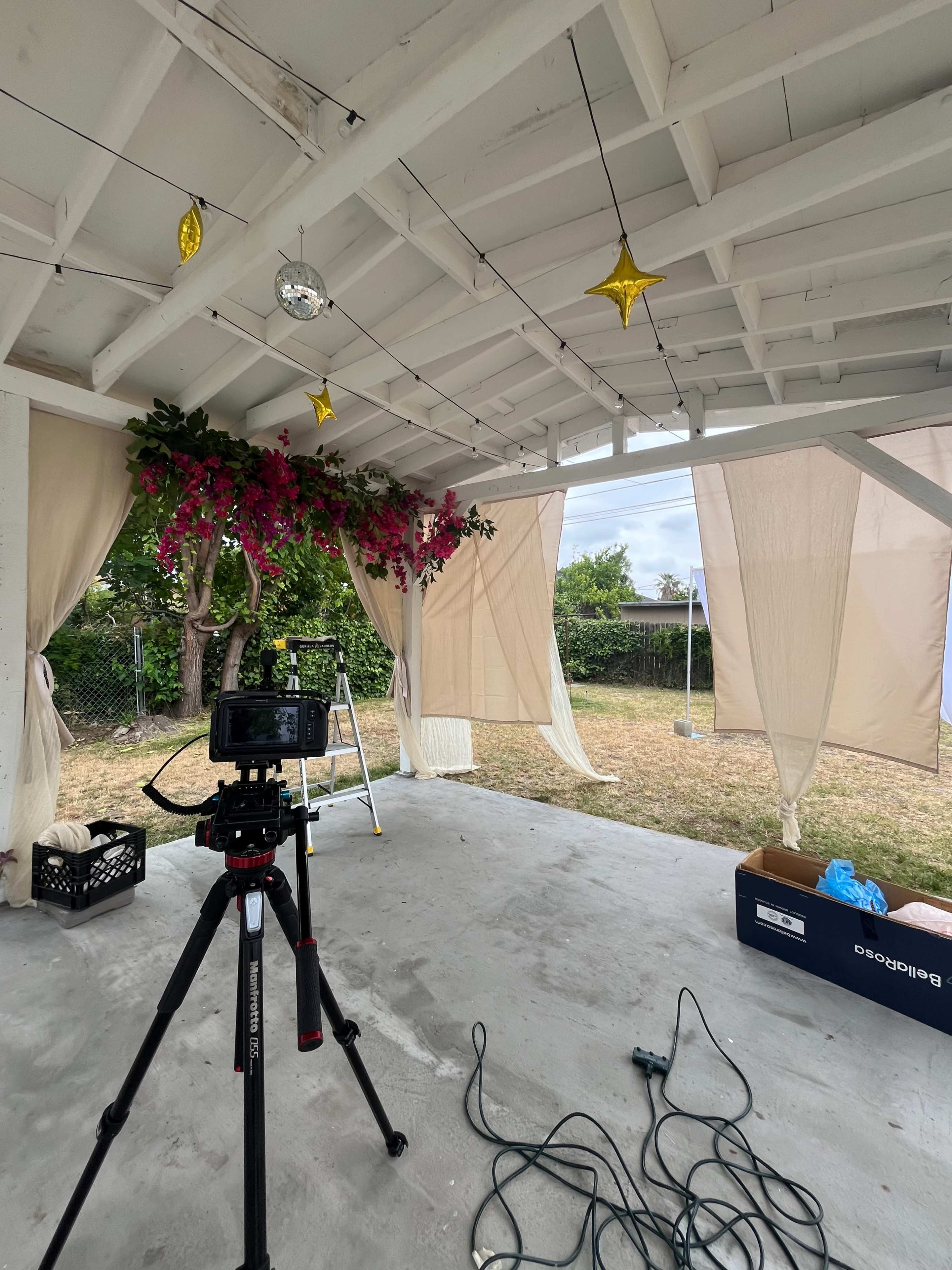 The image shows a covered outdoor space with a camera on a tripod, draped fabric, potted flowers, and decorative stars hanging from the ceiling.