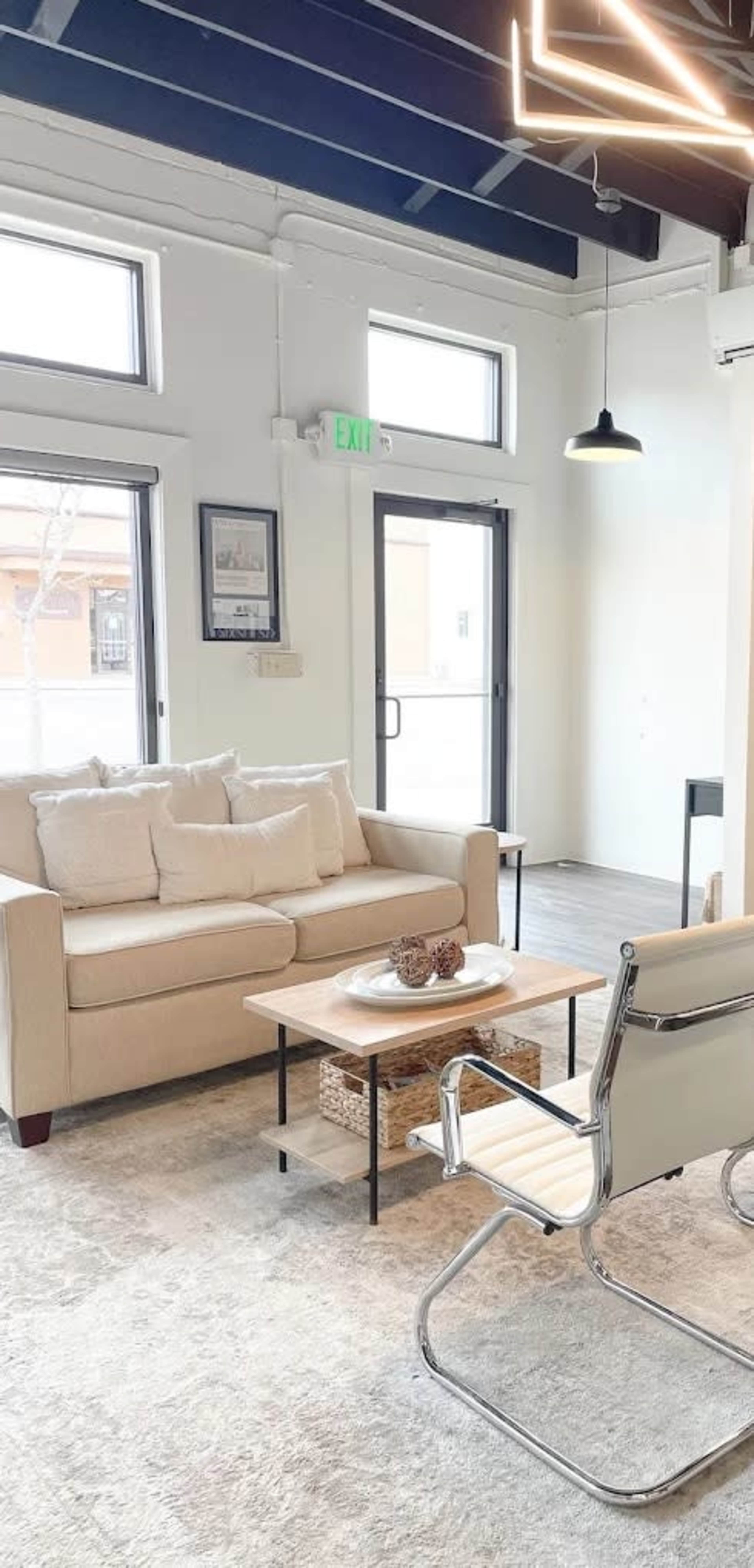 The image shows a modern living room with a beige sofa, a coffee table with decorative items, and a chair, set against a backdrop of large windows and industrial-style lighting.