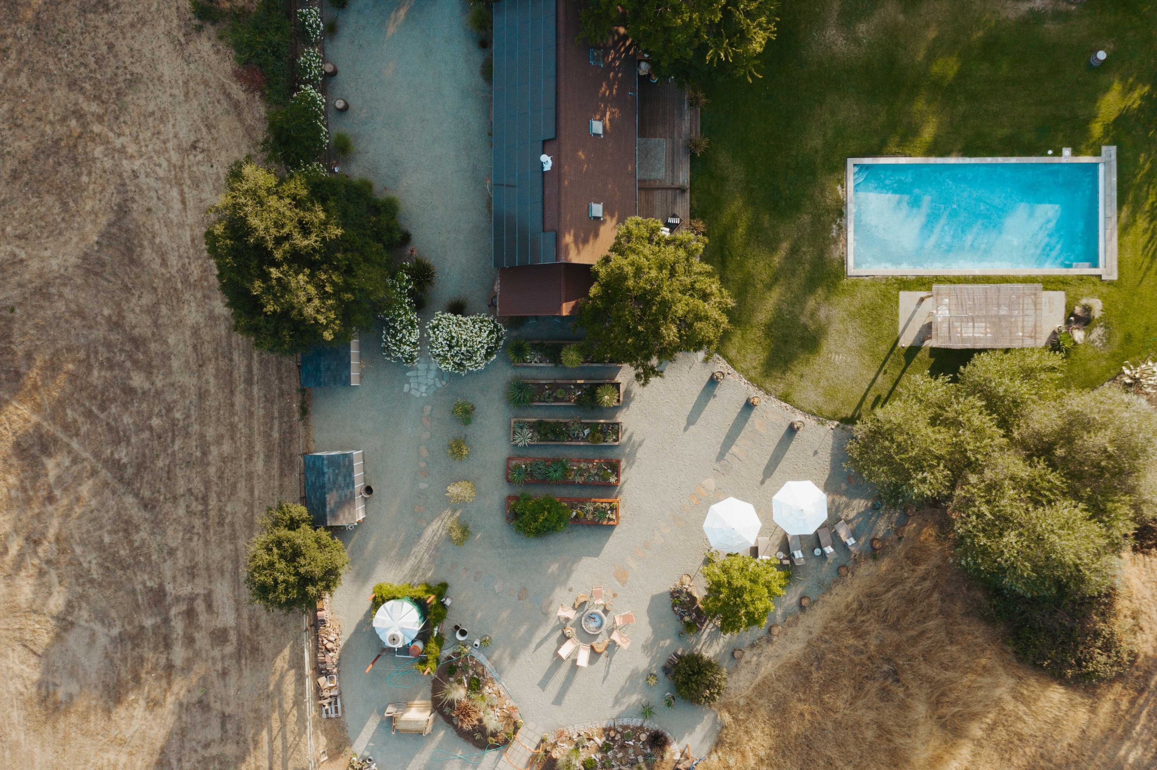 An aerial view shows a garden area with pathways, seating arrangements, a small swimming pool, and a house surrounded by trees and open fields.