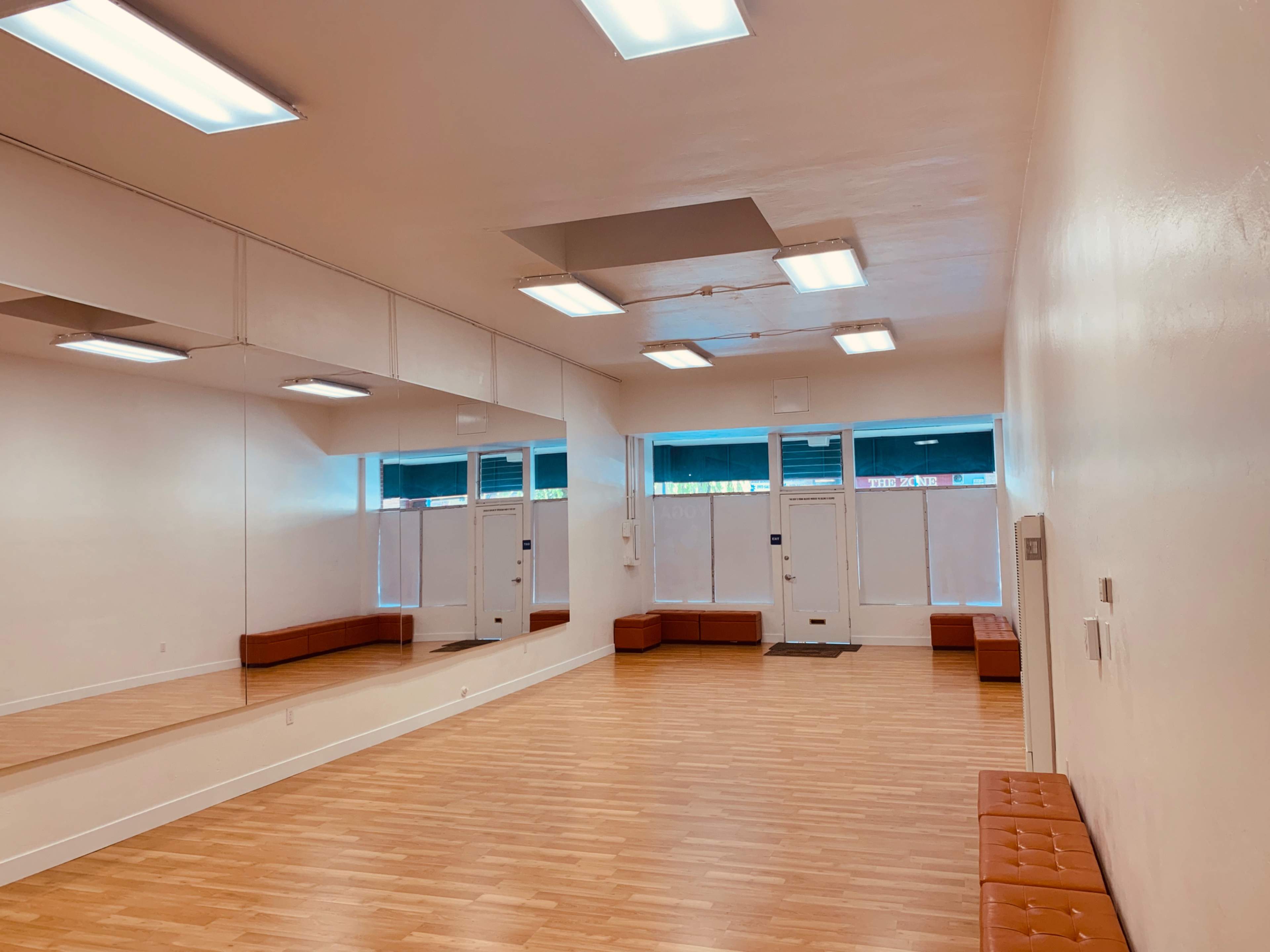 A spacious, empty room with wooden flooring, large mirrors on one wall, and two entrance doors at the front.