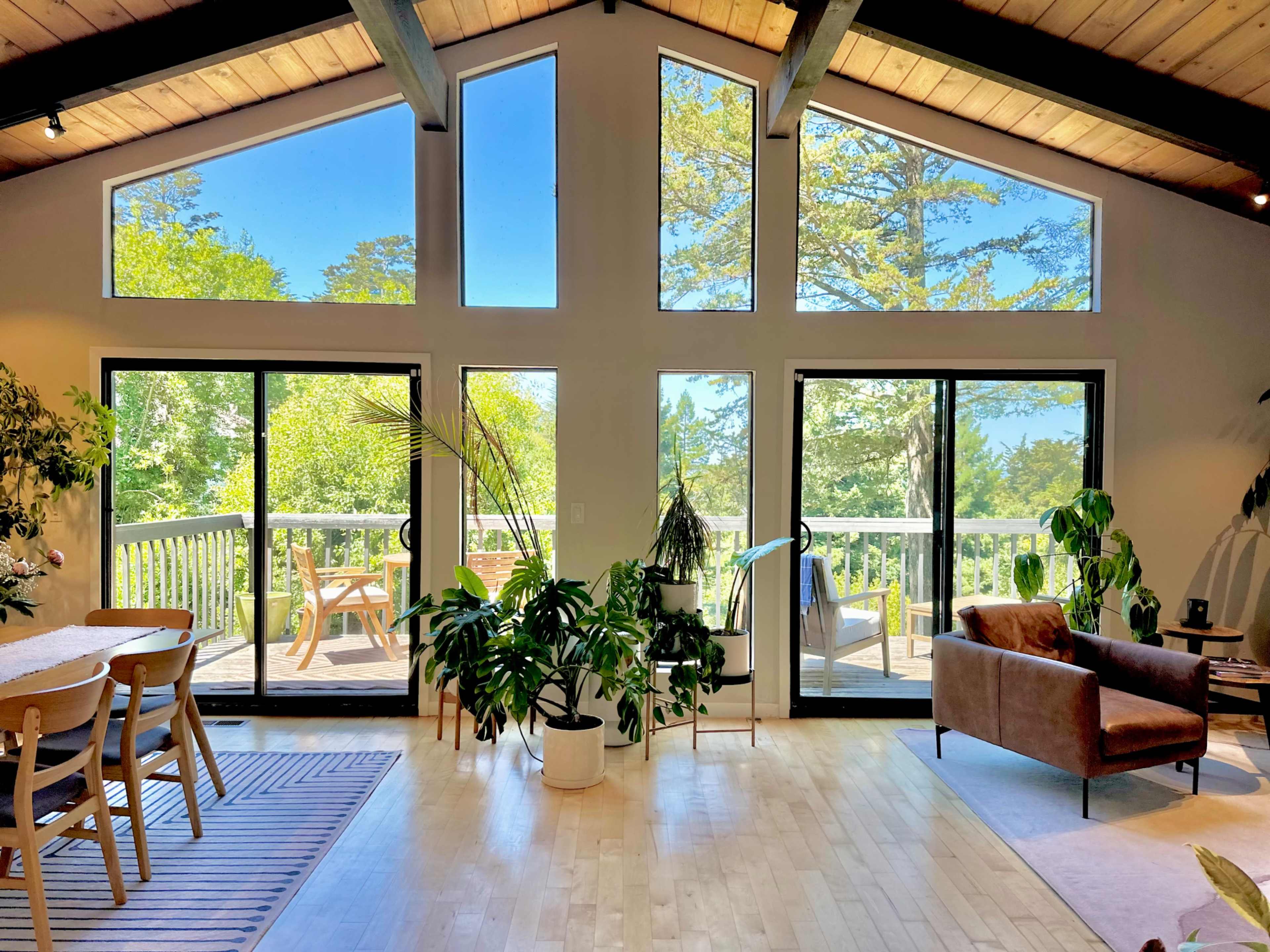 A bright, open interior space featuring large windows, a dining table, plants, and a view of a deck with outdoor seating.