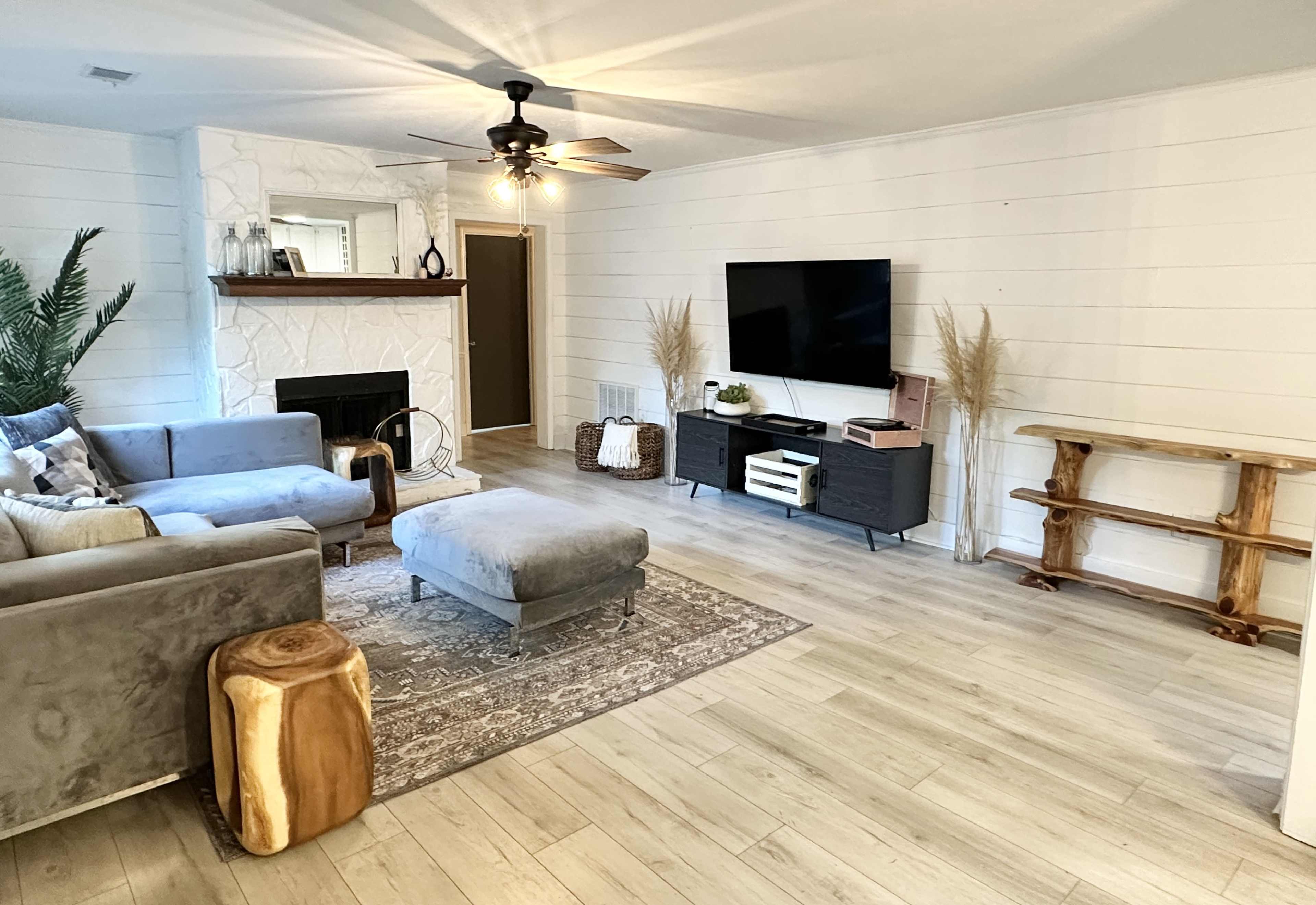 A spacious living room features a gray sectional sofa, a wooden coffee table, and a large TV mounted on a white wall with a fireplace and decorative plants.