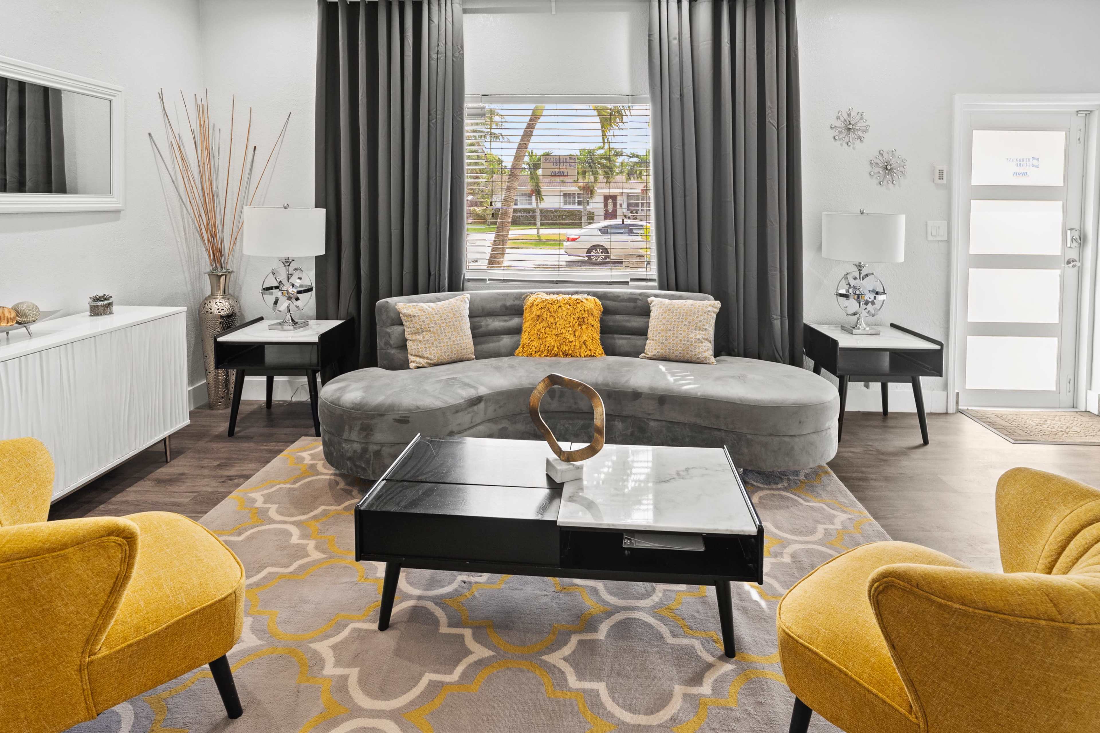The image shows a modern living room featuring a gray sectional sofa, yellow accent chairs, a black coffee table, and large windows with a view of the street outside.