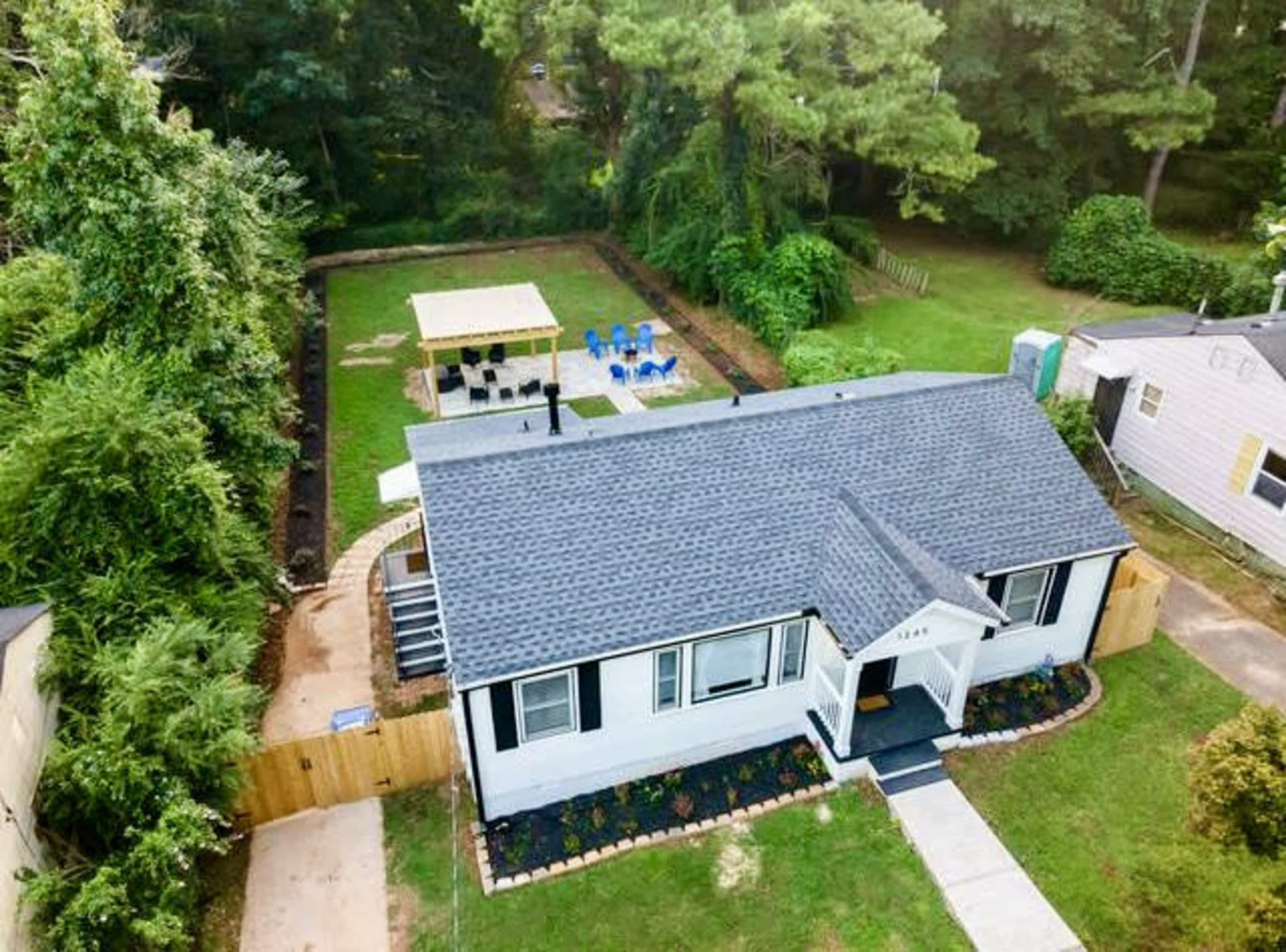 The image shows a single-story house with a black roof, surrounded by green foliage and featuring a backyard with a seating area and a small lawn.