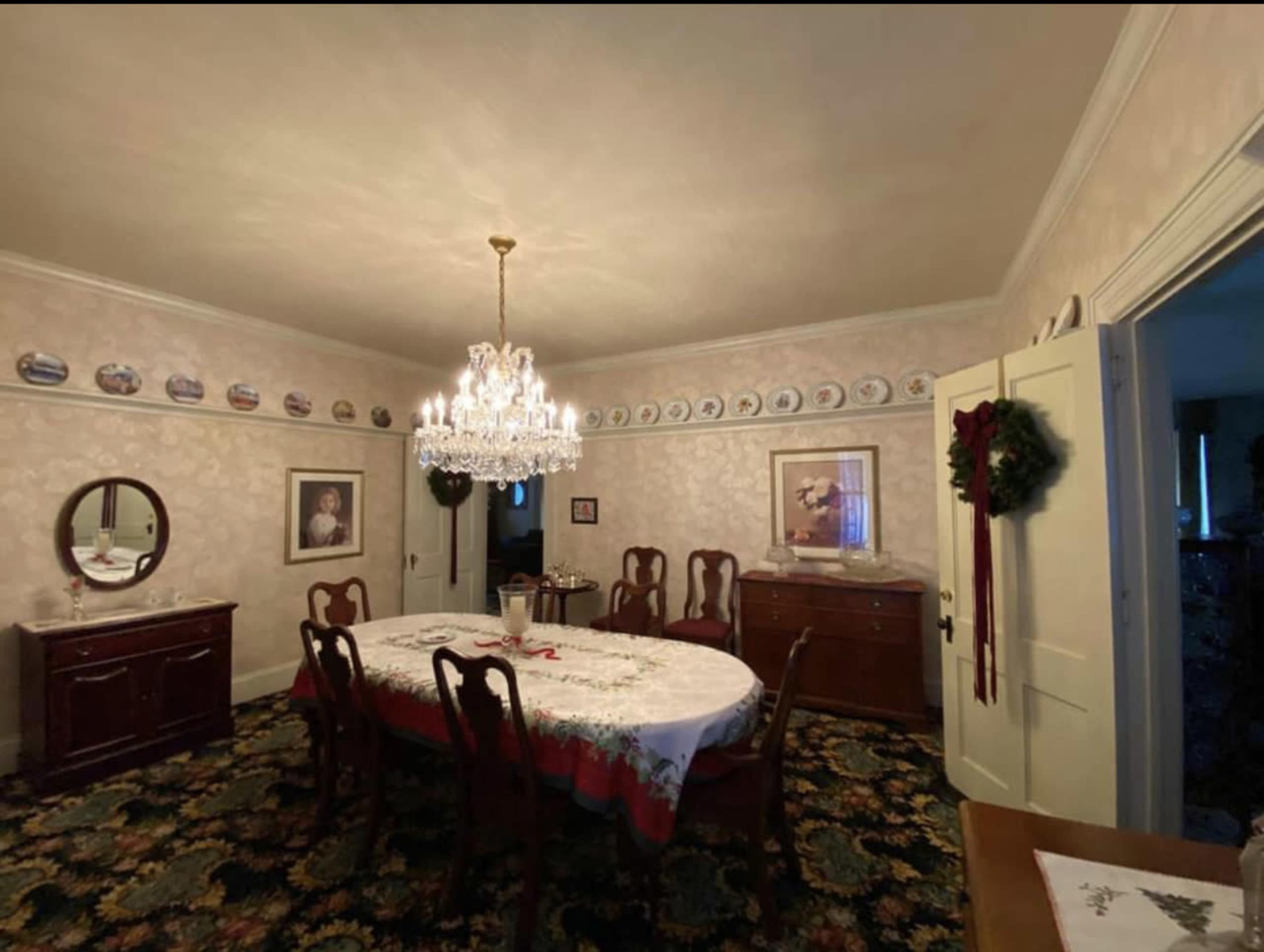 A dining room featuring a large table set with a tablecloth, surrounded by wooden chairs, a chandelier overhead, and decorated walls with plates and artwork.
