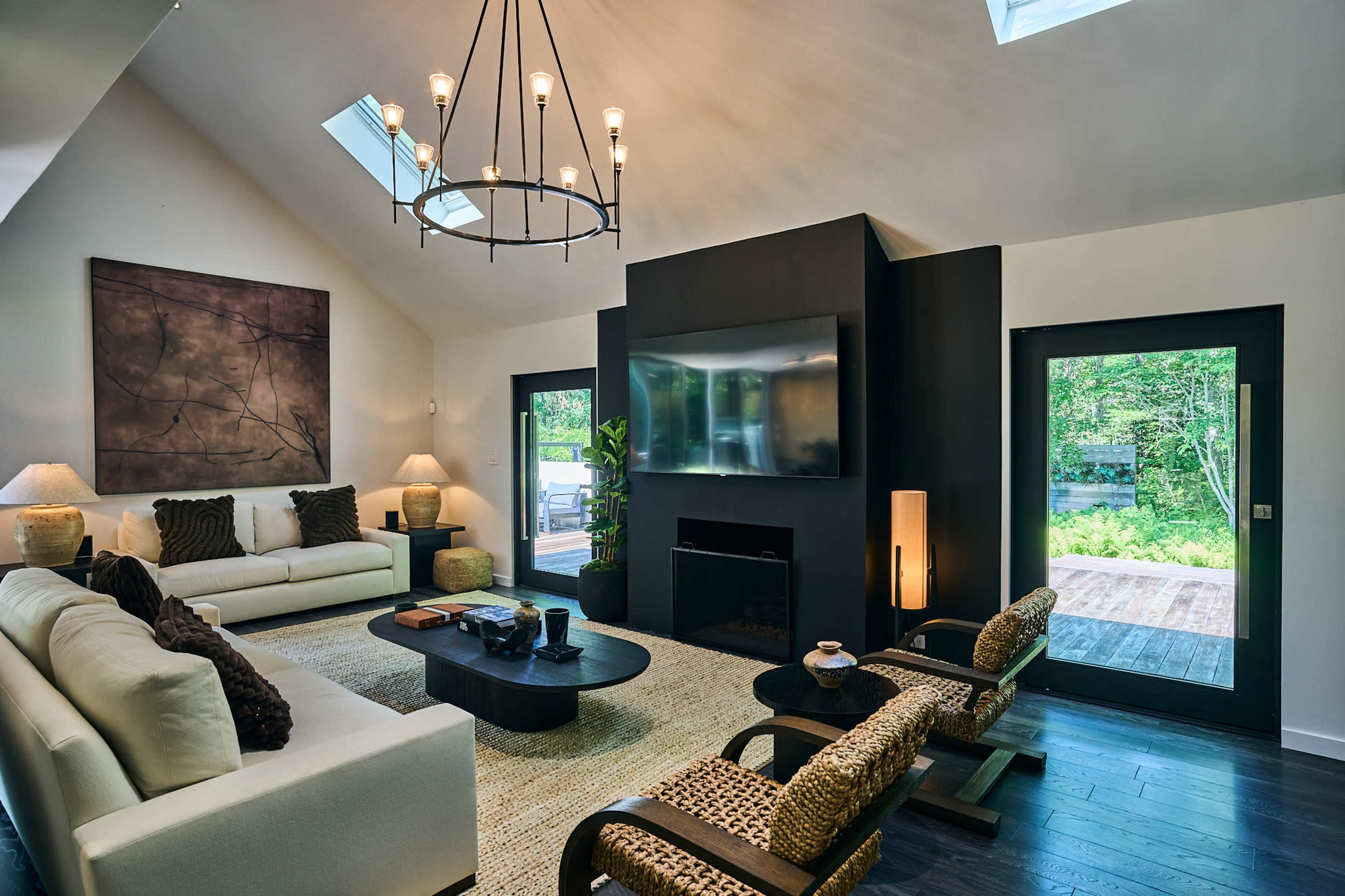 The image shows a modern living room with a large black TV mounted on a wall, a white sofa, wooden chairs, and large windows allowing natural light to enter.