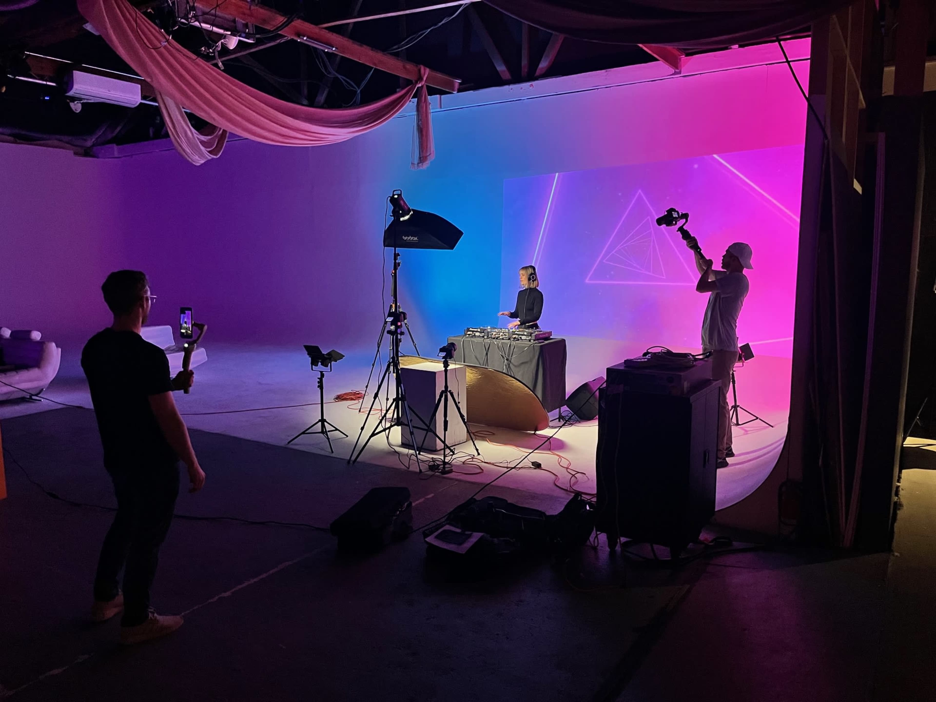 A person stands in a studio, holding a phone, while another person operates a camera in front of a DJ setup illuminated by colorful lights.