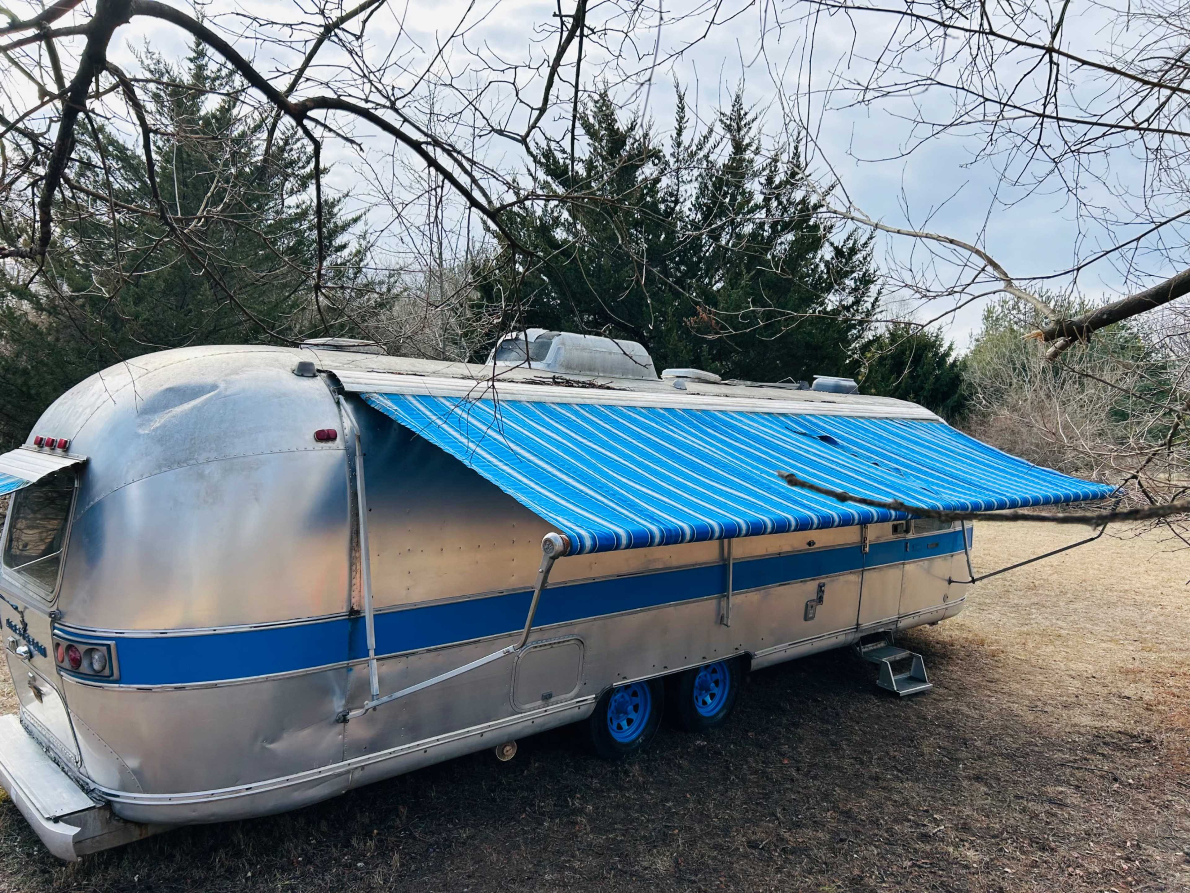 An Airstream trailer is parked on a grassy area, surrounded by trees, with a blue and white striped awning extended.