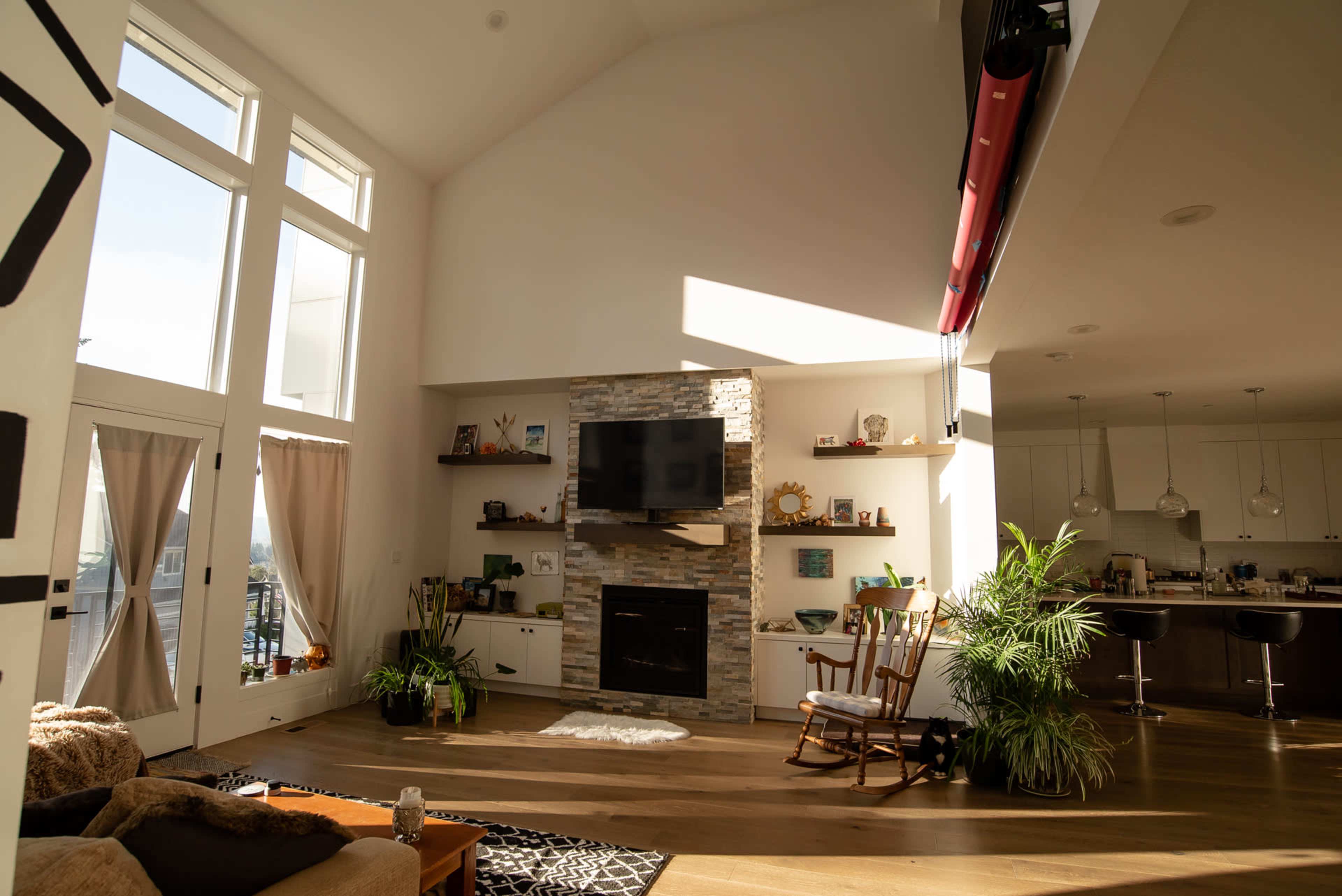 A bright interior features a stone fireplace with a mounted television, surrounded by shelves displaying decorative items, while a modern kitchen is visible in the background.