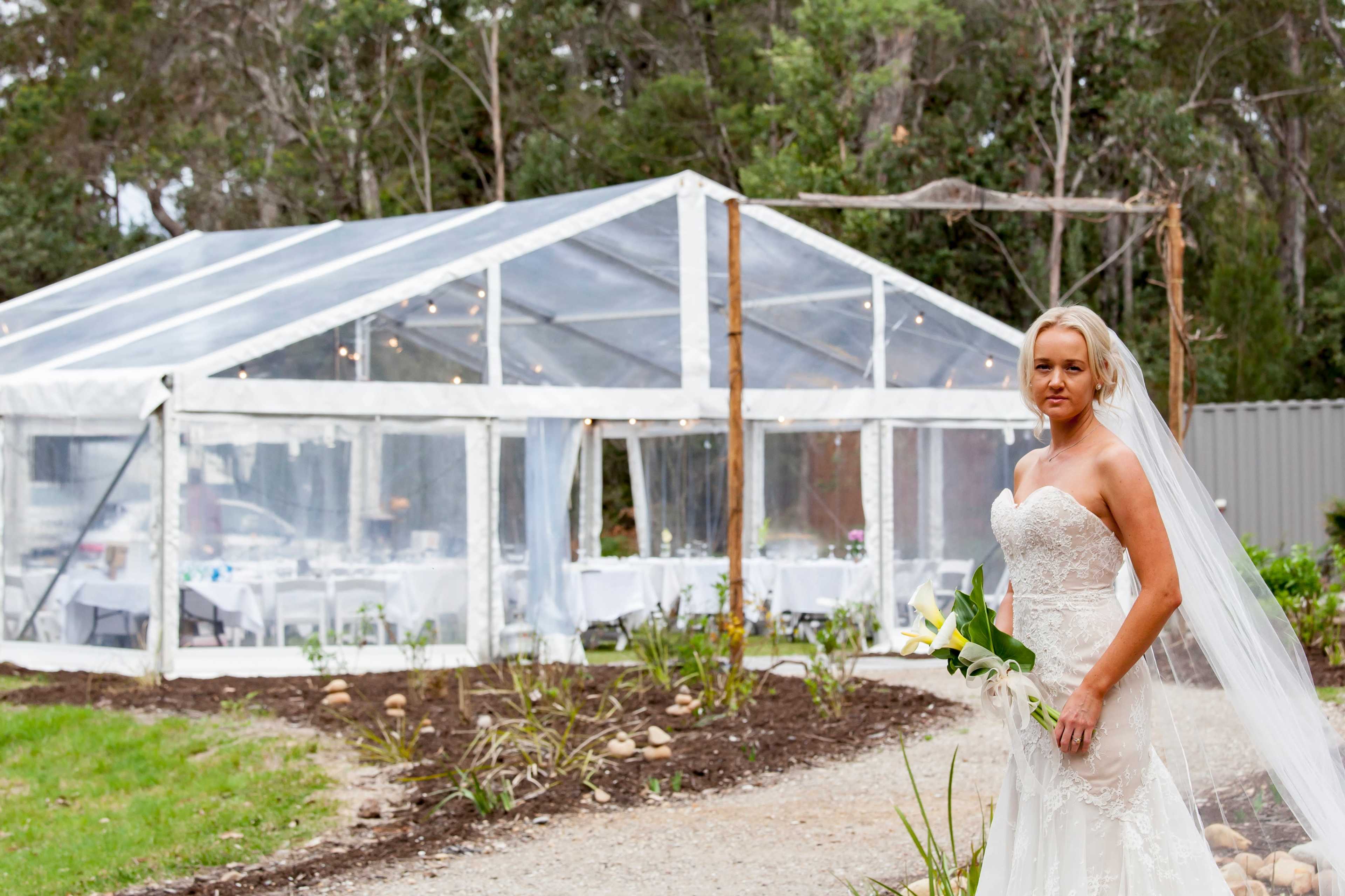 A bride in a white gown holds a bouquet of lilies while standing in front of a transparent tent set up for a wedding reception.