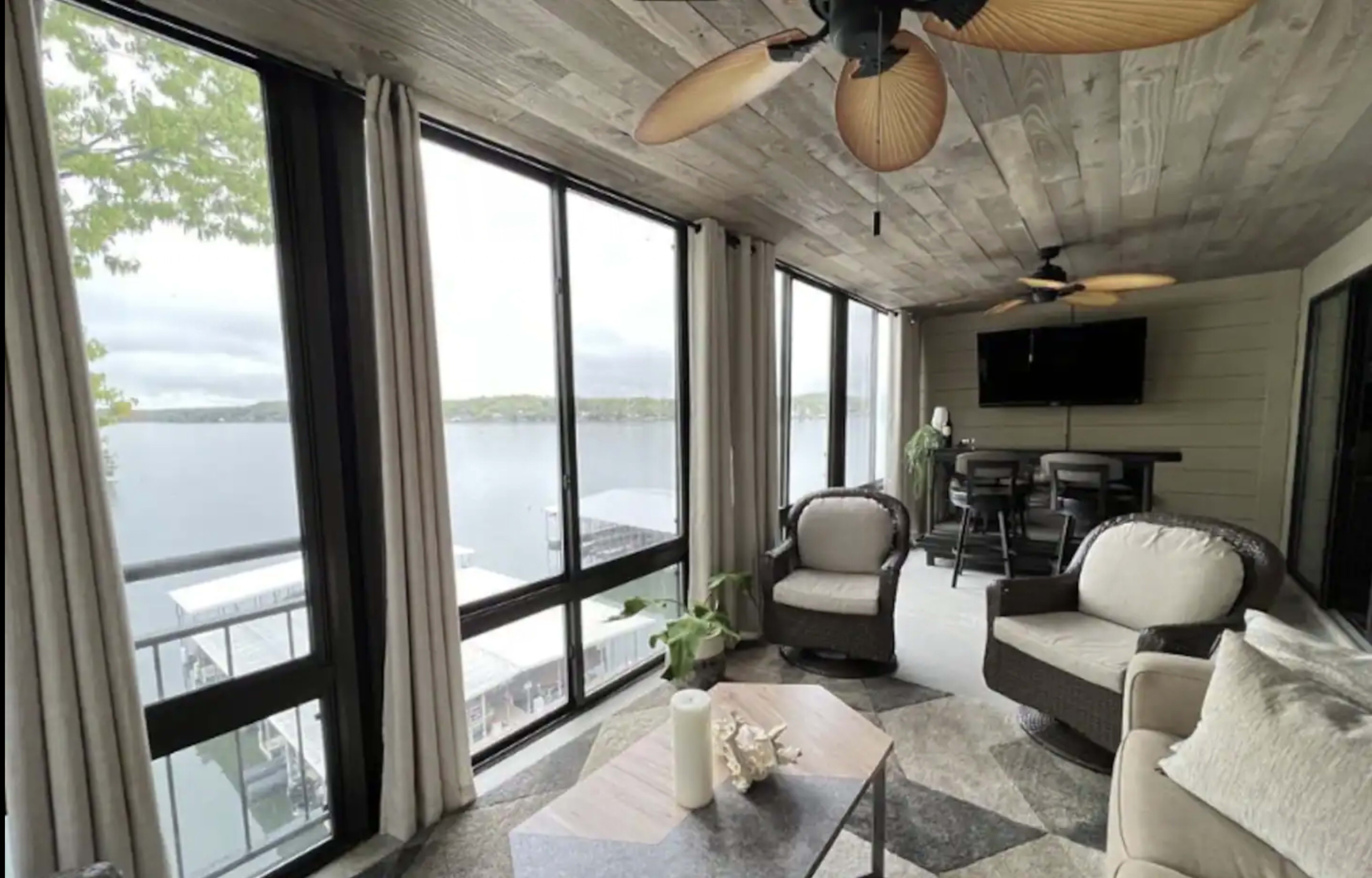 A sunroom with large windows overlooking a body of water, featuring comfortable seating, a coffee table, and a ceiling fan.