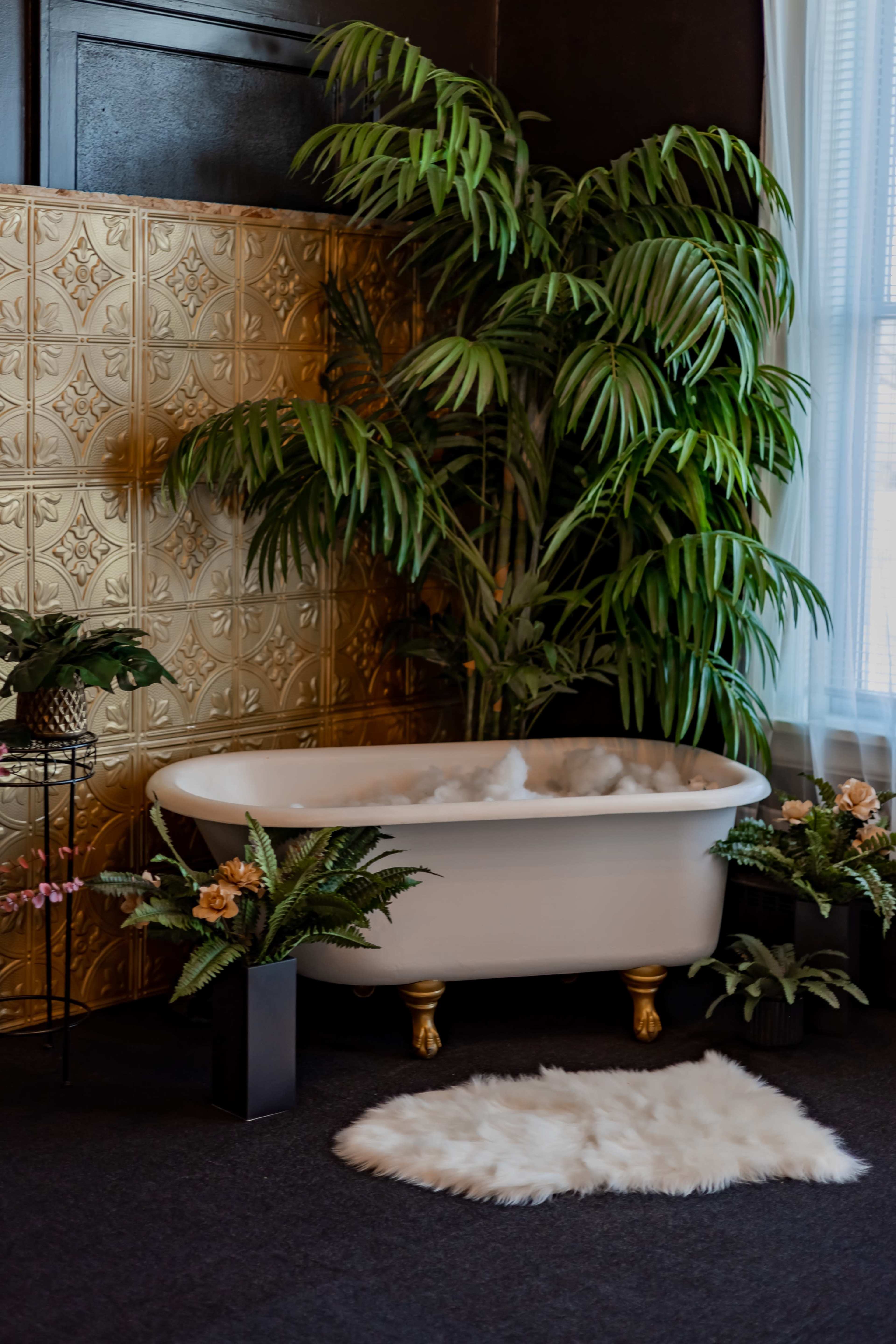 A freestanding white bathtub is surrounded by potted plants and decorative flowers against a gold-patterned wall.