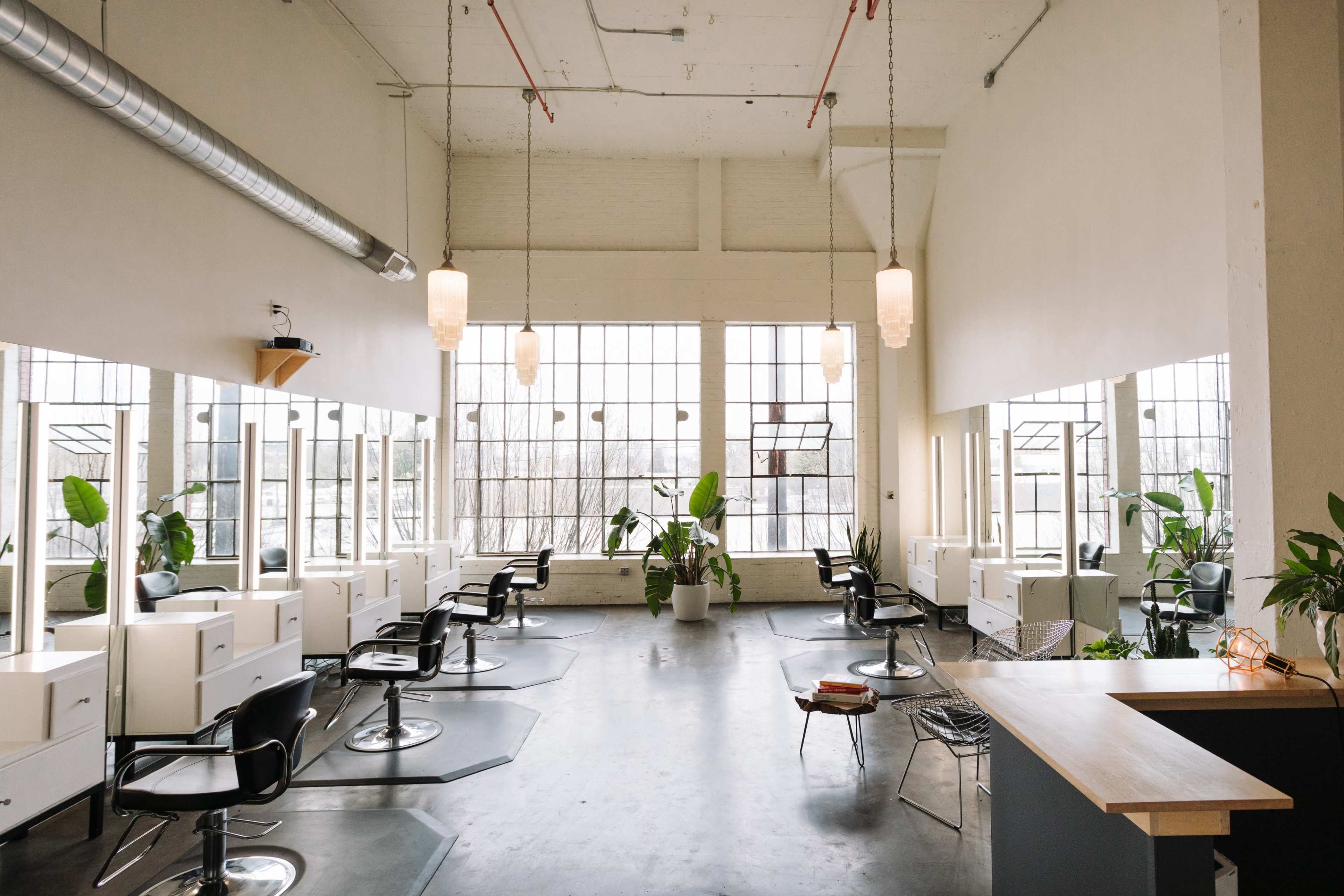 A spacious hair salon with several styling stations, large windows, and decorative plants.
