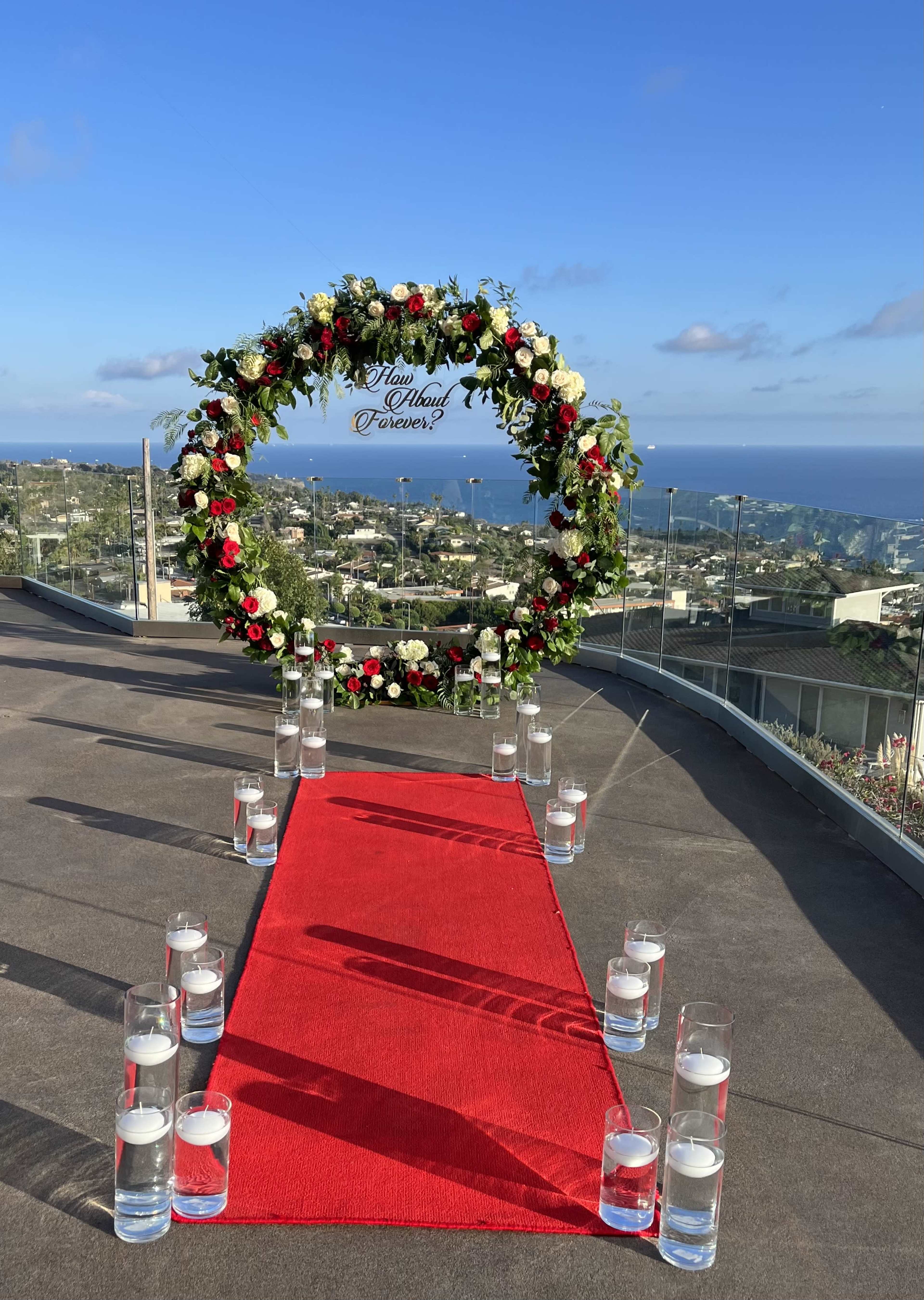 A circular floral arch adorned with red and white flowers stands at the end of a red carpet, flanked by glass candles, overlooking a panoramic view of a coastal landscape.