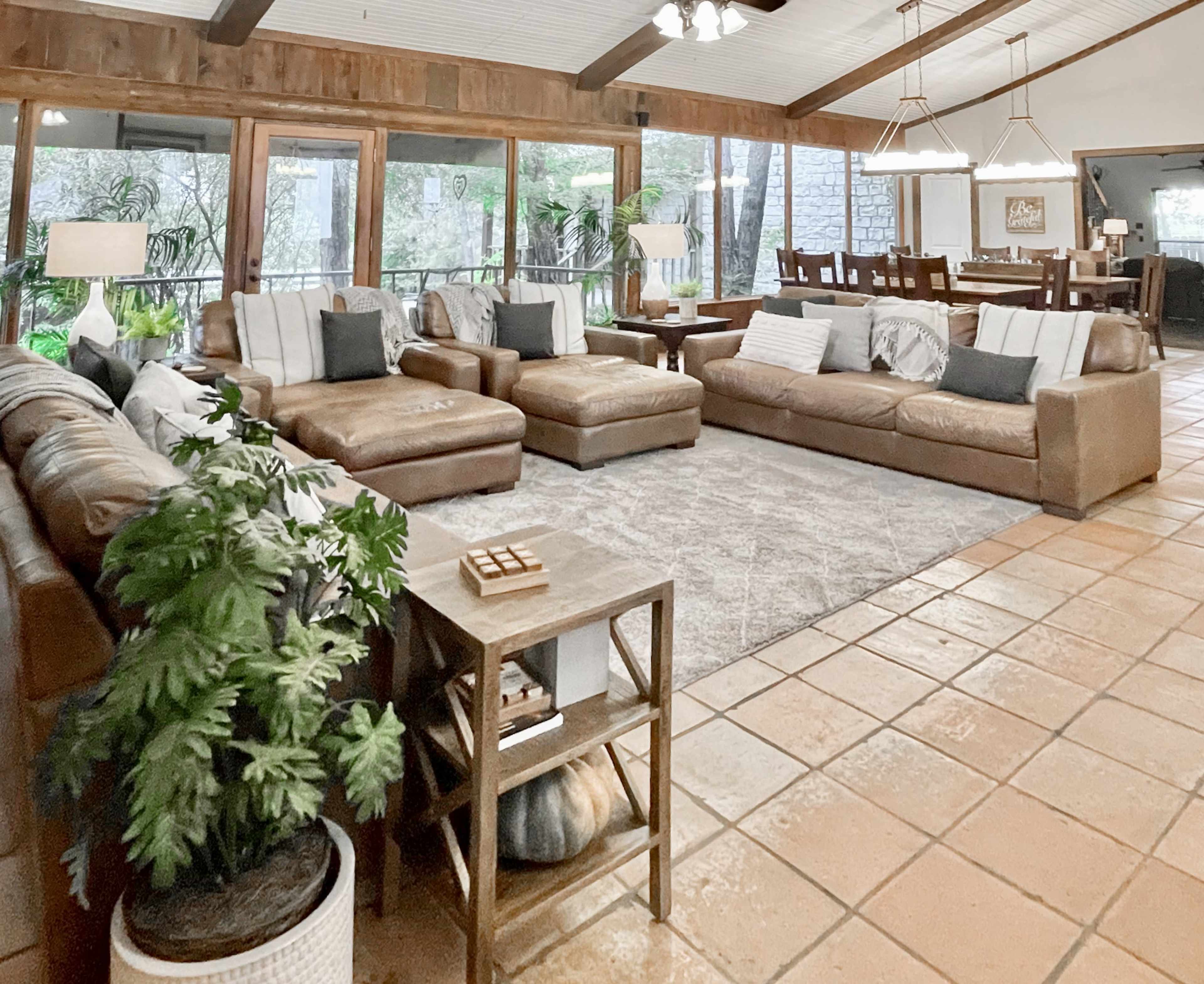 A spacious living area with leather sofas arranged around a large rug, accompanied by a dining table in the background and large windows letting in natural light.
